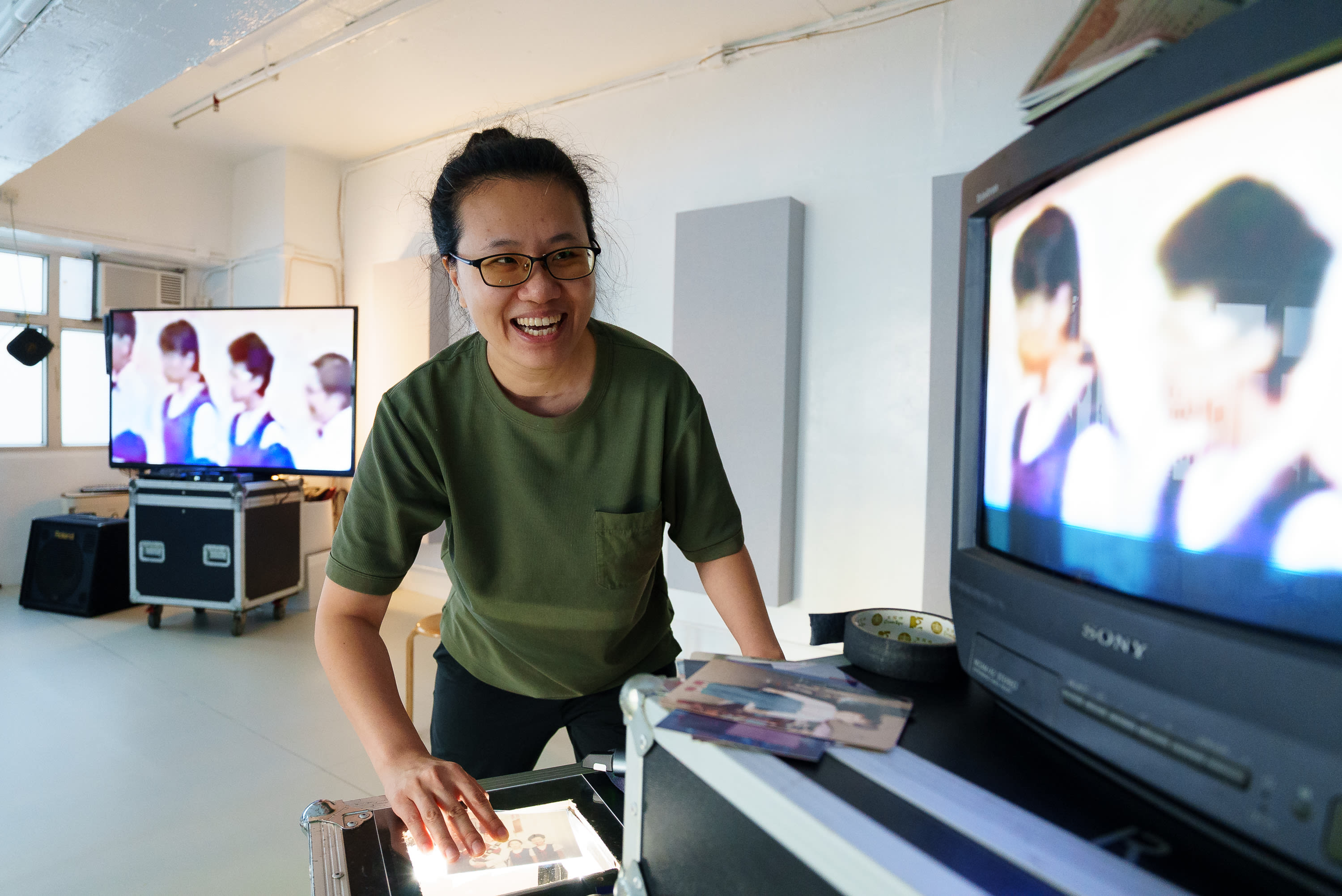 在明亮的排練室中，演員李婉晶（Michelle Li）正於燈箱前整理舊照片，為天台製作的當代劇場作品《卡啦OK》進行排練。背景的螢幕展示著製作過程中的影像記錄。「《卡啦OK》排練」