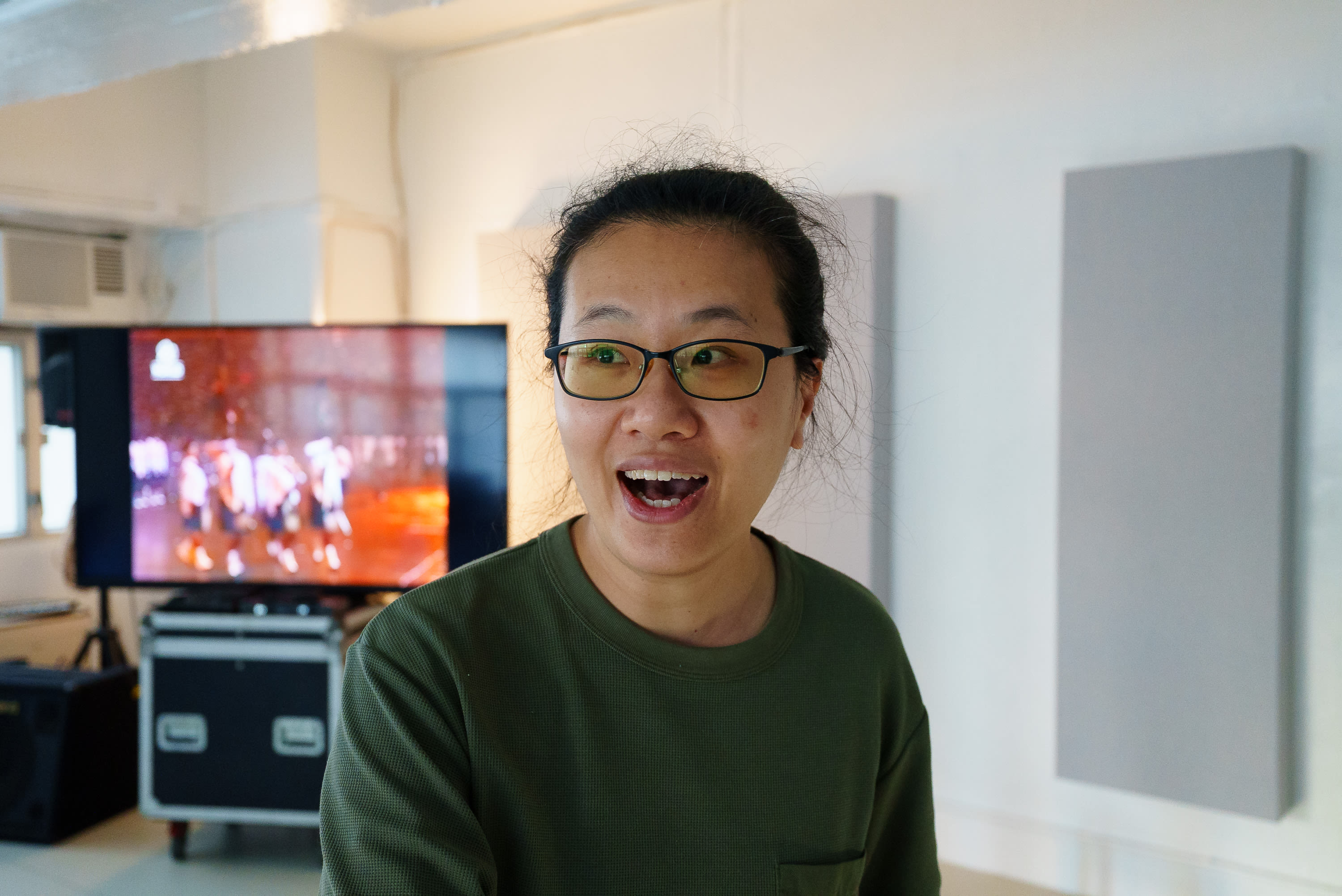 天台製作（Rooftop Productions）當代劇場作品《卡啦OK》排練現場，演員李婉晶神情投入且充滿活力。她戴著眼鏡，身穿綠色長袖上衣，背景可見舞台設備及播放著演出畫面的螢幕。《卡啦OK》排練。