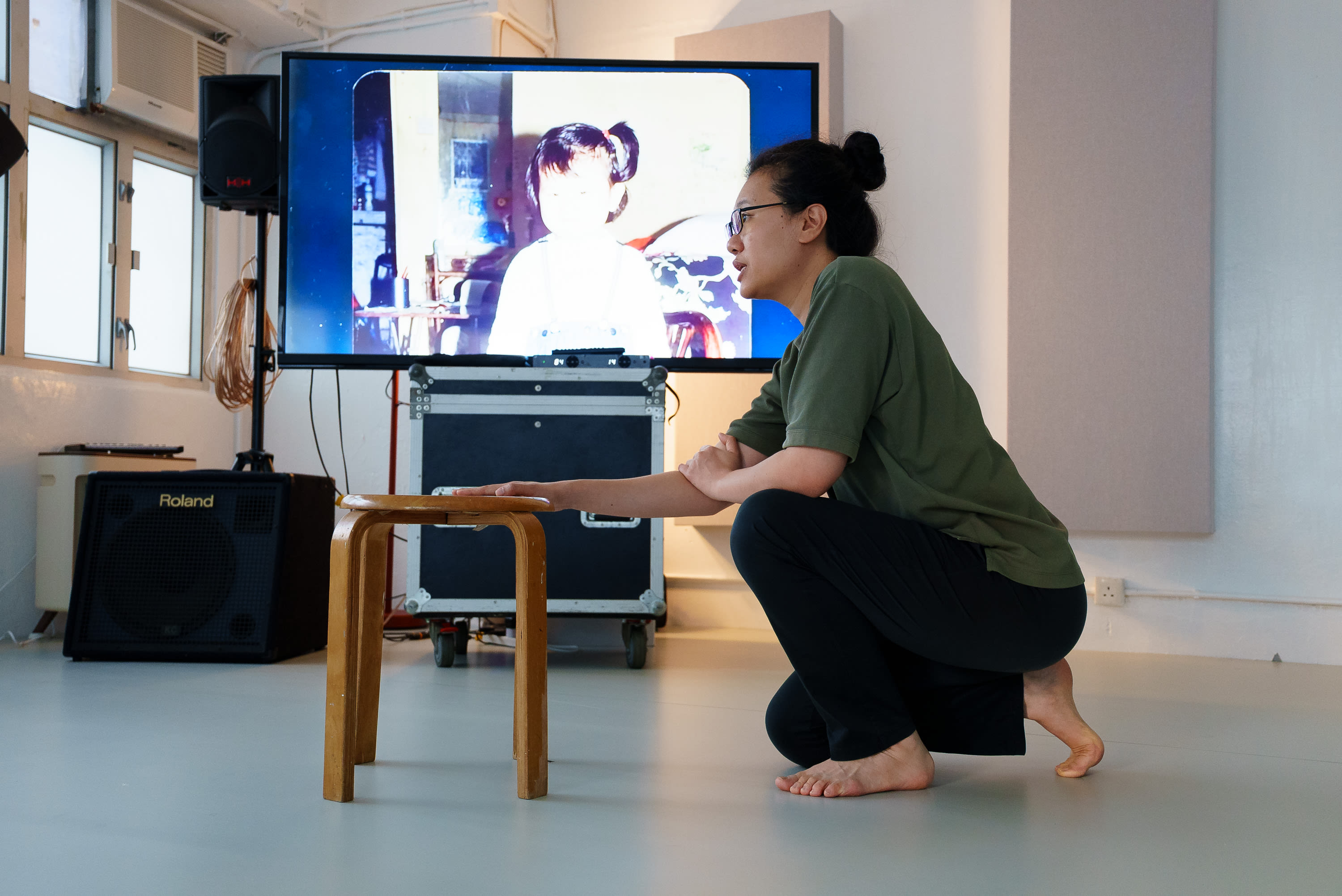 In a rehearsal for the contemporary Hong Kong theatre production 'Karaoke' (《卡啦OK》), actor Michelle Li is captured in a poignant moment on stage. Dressed in comfortable rehearsal attire, she kneels on the floor beside a simple wooden stool, her hand resting thoughtfully on its surface. Behind her, a large screen displays a nostalgic, stylized digital image of a young girl, emphasizing the themes of memory and performance inherent in Rooftop Productions' (天台製作) work. The studio setting is minimalist, lit by professional stage equipment. Karaoke rehearsals.
