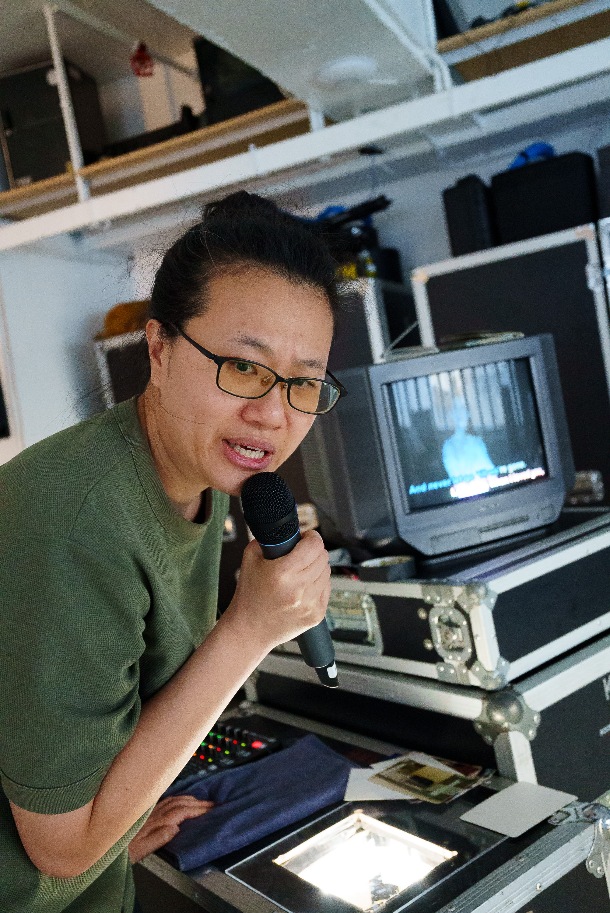 Performer Michelle Li holds a microphone and rehearses for the Rooftop Productions (天台製作) contemporary theatre piece 'Karaoke' (《卡啦OK》). She stands behind a professional technical setup featuring a vintage CRT monitor displaying song lyrics, capturing the immersive, nostalgic atmosphere typical of this Hong Kong stage production. 'Karaoke rehearsals'