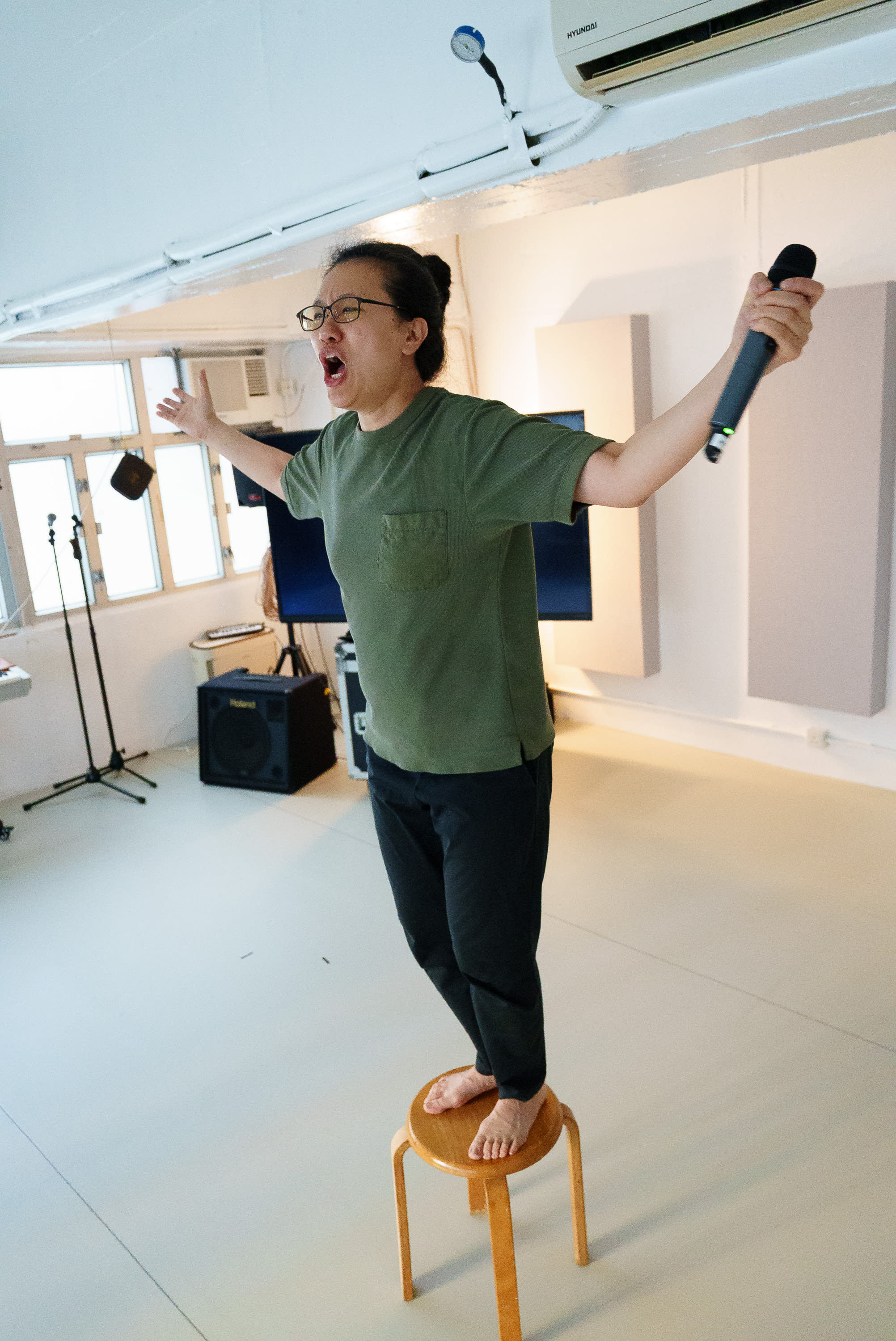 演員李婉晶赤腳站在木製圓凳上，雙臂向兩側伸展，一手持麥克風，頭部後仰，以充滿戲劇張力的姿態投入於天台製作（Rooftop Productions）當代劇場作品《卡啦OK》的排練之中。排練室明亮的佈光凸顯出這場《卡啦OK》排練現場那份原始而純粹的戲劇張力。