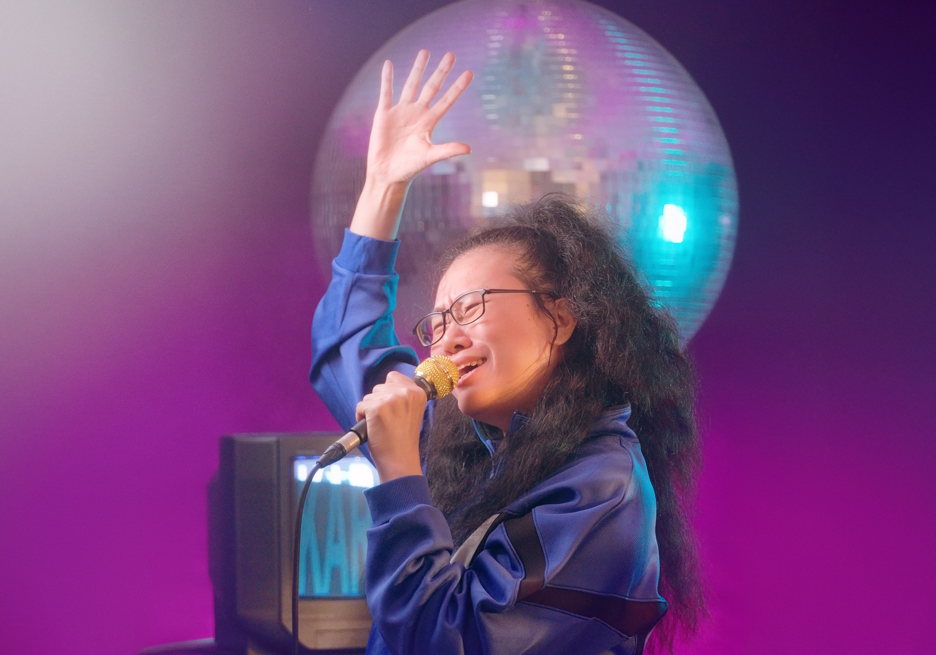 Michelle Li 在天台製作的當代劇場演出中，深情投入地手持麥克風演唱。她身穿休閒藍色運動套裝，神情專注激昂，一手舉向半空，背景映襯著巨大的閃爍迪斯可球與一台復古方型電視機，充滿香港當代舞台表演的懷舊氛圍。「Michelle Li - Karaoke」