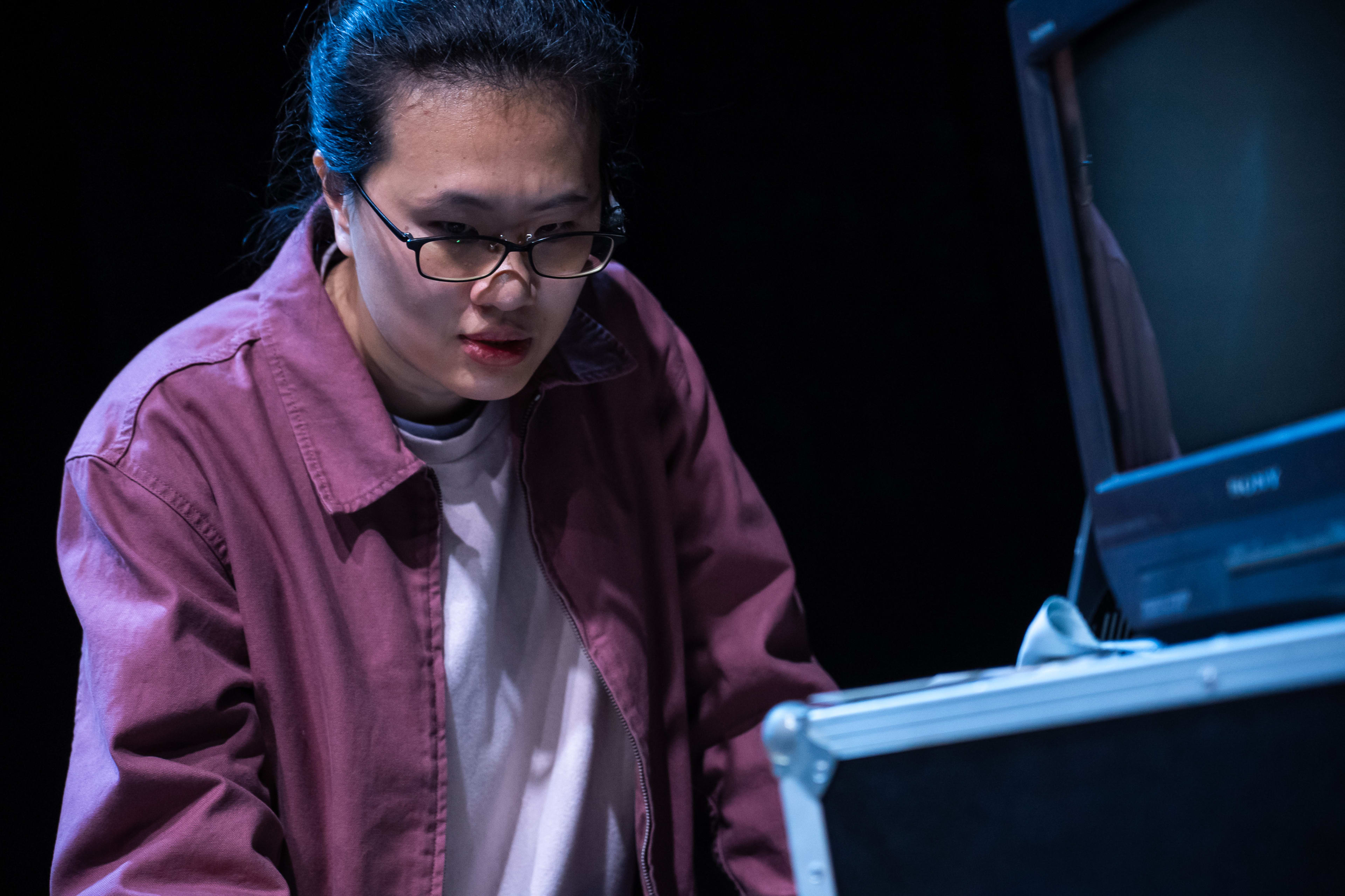 在天台製作（Rooftop Productions）當代劇場作品《卡啦OK》的排練場中，演員李婉晶站在昏暗的舞台燈光下，神情凝重且若有所思地望向眼前的舊式錄影機與螢幕，呈現出戲劇性的冷調氛圍。這場演出深入探討記憶與失智症等議題，李婉晶透過台詞演繹道：「阿茲海默症……有聽過，但實質是甚麼？」