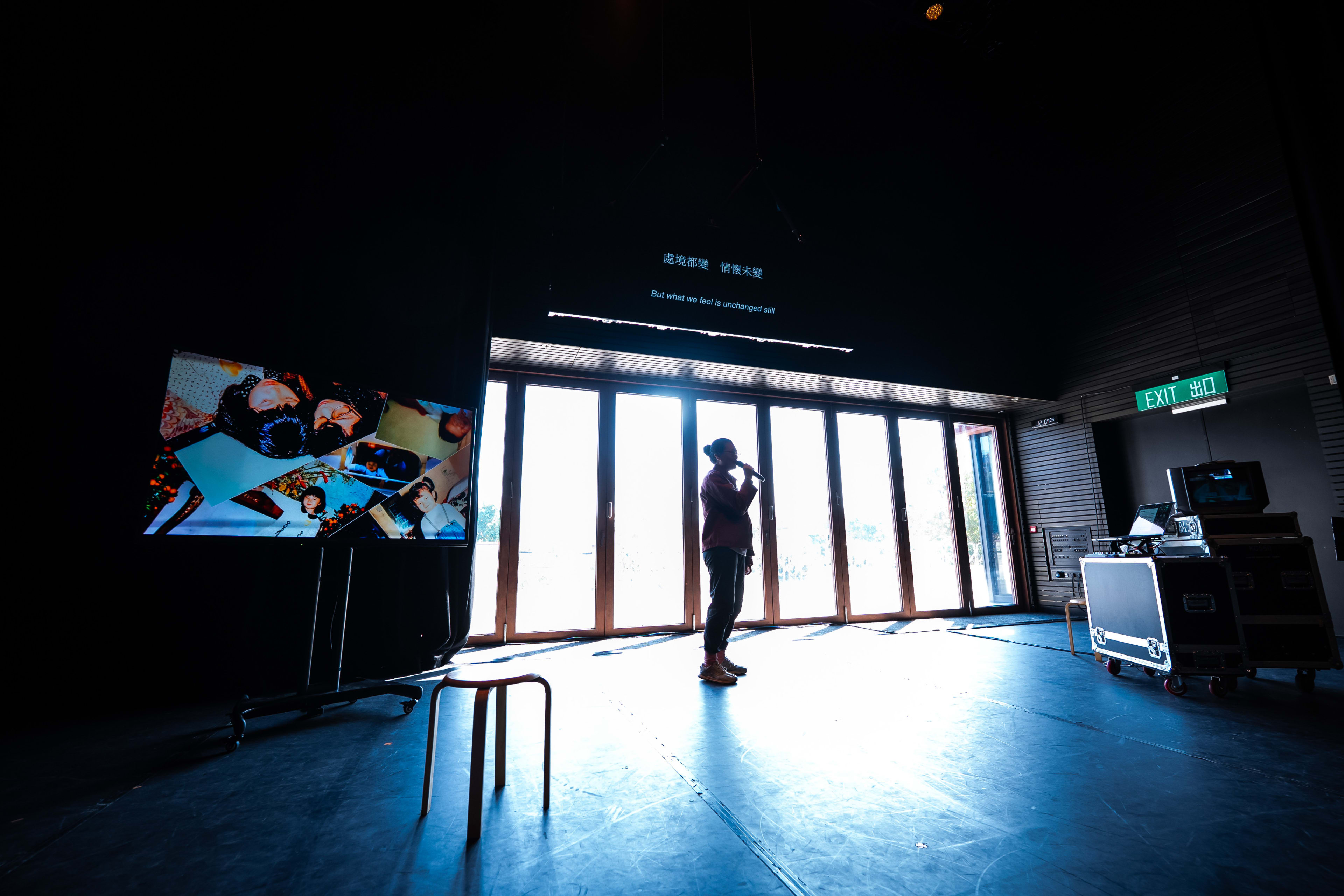 In a dimly lit stage space for Rooftop Productions' contemporary theatre piece 'Karaoke', actor Michelle Li stands in silhouette against a brightly backlit glass wall, holding a microphone. Beside her, a large screen displays a collage of intimate photographs, while a stool stands empty in the foreground, evoking a sense of solitary reflection typical of experimental Hong Kong stage design. As she stands in the spotlight, she captures the essence of the play’s theme: "But what we feel is unchanged still."