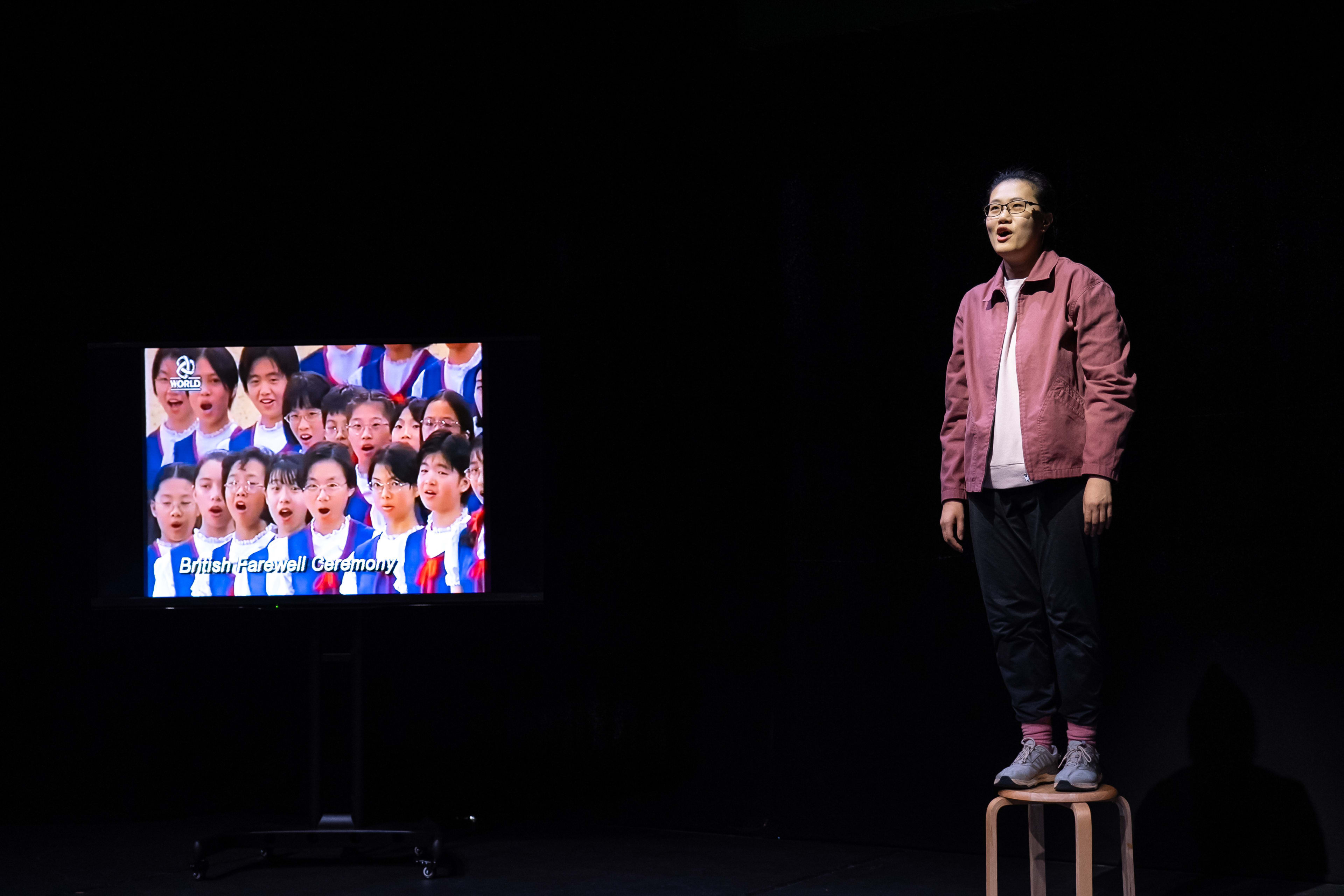 在天台製作的當代劇場作品《卡啦OK》的極簡舞台上，演員李婉晶站在一張小凳子上。她身穿休閒的紫紅色外套並戴著眼鏡，表情投入地進行演出，眼神略帶仰望。舞台左側的顯示屏播放著一張張年輕臉孔的合照，營造出充滿懷舊感的檔案影像。這一幕展現了該劇對記憶與身份議題的深刻探討。李婉晶唱道：「世上所有的兒童。」