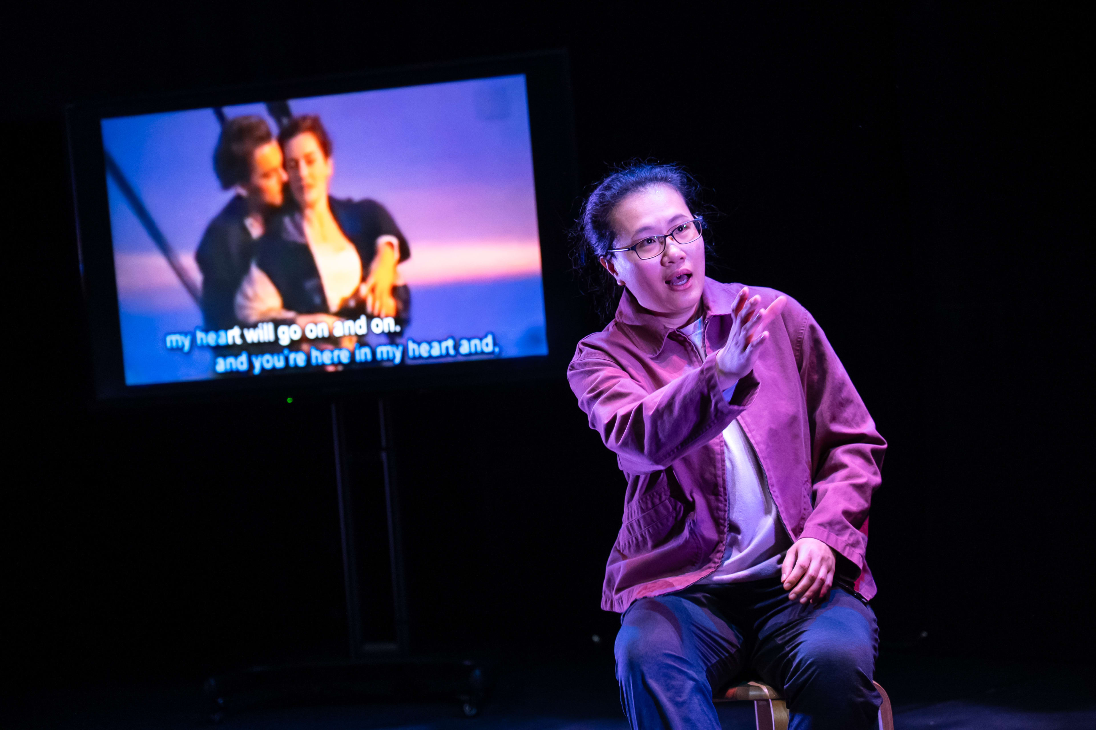 在天台製作的當代香港劇場作品《卡啦OK》中，演員李婉晶坐在舞台中央的凳子上，身處強烈的舞台燈光與全黑背景之間。她伸出手臂，表情流露出深沉而細膩的情感。身後的螢幕正播放電影《鐵達尼號》的經典浪漫劇照，畫面下方的歌詞字幕與舞台氛圍相互映襯。她深情演繹道：「不管是遠是近，無論你在哪裏。」