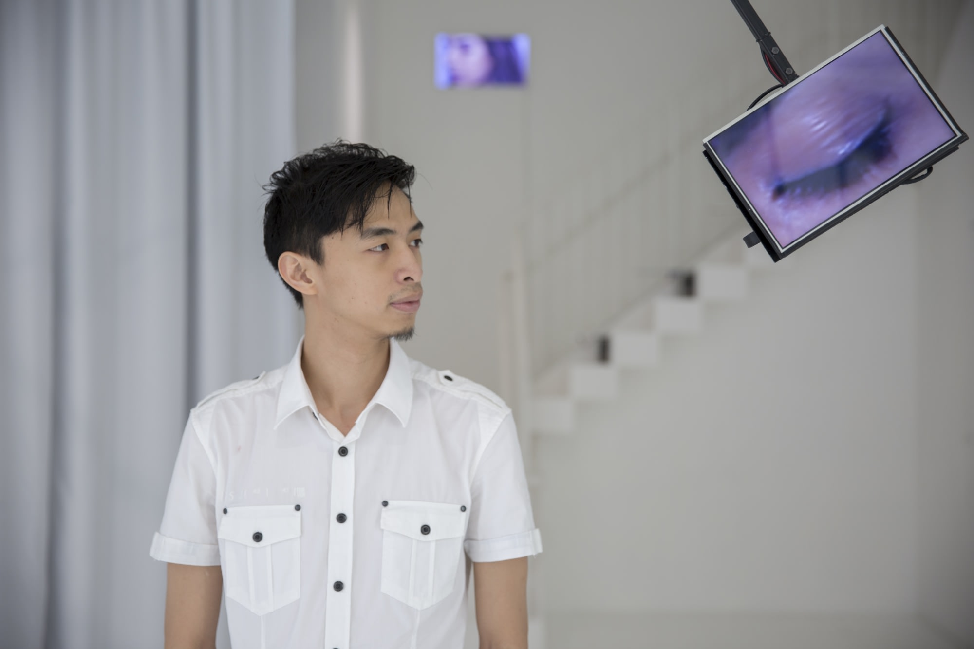 天台製作 (Rooftop Productions) 舞台演出宣傳照，由香港演員黃智銓擔任主角。他身穿白色恤衫，神情專注地看向右方，該處懸掛著一個顯示人類眼睛特寫投影的螢幕。簡約的單色調場景呈現出香港當代實驗劇場的獨特美學。劇照。