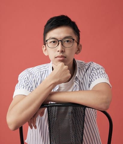 香港演員盧宜敬（Kingston Lo）的個人劇照，背景為鮮明的紅色，展現當代香港劇場的簡約美學。他坐在黑色網狀椅背的椅子上，雙臂交疊靠在椅背，並將下巴輕靠在手上，神情沉穩思索。他身穿白色直條紋短袖襯衫內搭白色T恤。一張定裝照。