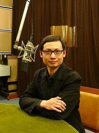 香港資深劇評人及天台製作（Rooftop Productions）藝術工作者曲飛的錄音室頭像照。他身穿黑色襯衫，雙手交叉置於綠色桌面上，背景為專業吊掛式電容麥克風與棕色布幕。這是一張人物頭像照。