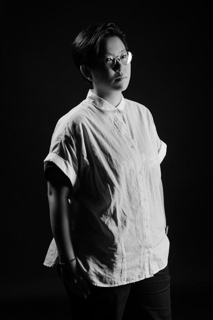 天台製作（Rooftop Productions）劇團演員賴嘉琪的黑白宣傳照。照片中，她佩戴眼鏡，身穿短袖翻領襯衫，在強烈對比的舞台燈光與純黑背景下，展現出凝神思考的神情。宣傳照。