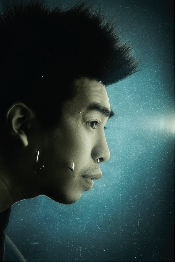 演員吳紹熙在水中拍攝的戲劇性側面特寫，展現出天台製作 (Rooftop Productions) 當代香港劇場表演中常見的沉思氛圍。光影效果夢幻且帶有冷色調，氣泡環繞在他的臉龐周圍，凸顯出強烈的電影美學。定裝照。