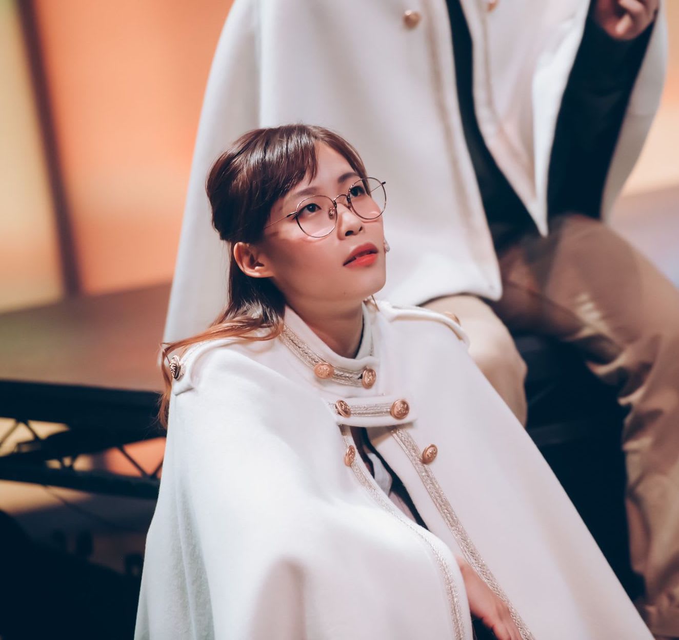 天台製作（Rooftop Productions）當代劇場演出的劇照，演員劉嘉盈身穿帶有金色鈕扣與精緻飾邊的白色軍裝風格披風，眼神專注地仰望上方，表情深沉而富有戲劇張力。柔和的舞台燈光突顯了戲服與眼鏡的細節，呈現出香港獨立劇場的高水準製作質感。頭像攝影。