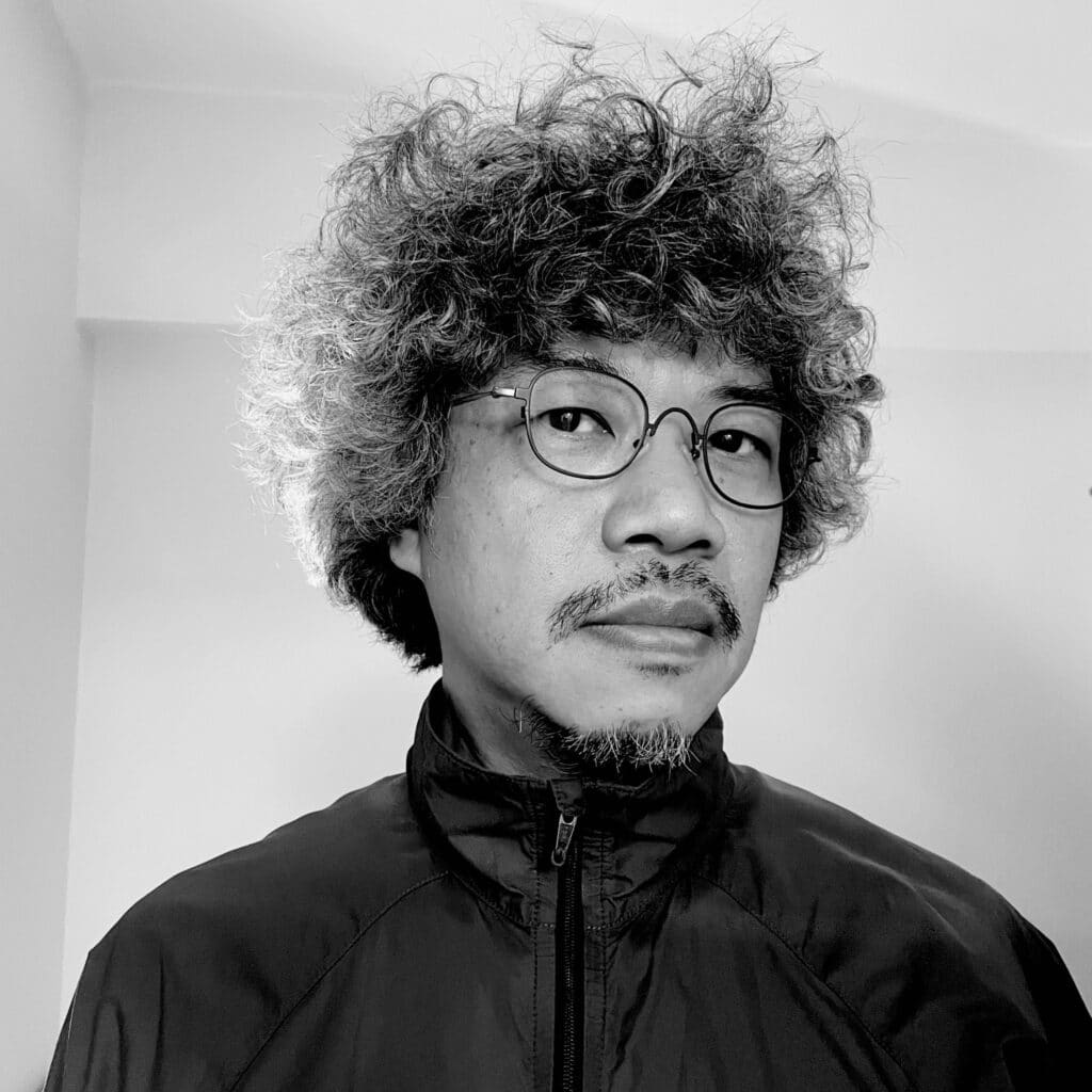 香港藝術家利志達的黑白頭像，他曾參與當代劇場及跨界藝術創作。照片中的他戴著圓框眼鏡，身穿拉鍊外套，神情沉穩，展現出天台製作（Rooftop Productions）實驗性舞台藝術作品中所蘊含的深刻人文氣息。頭像照。