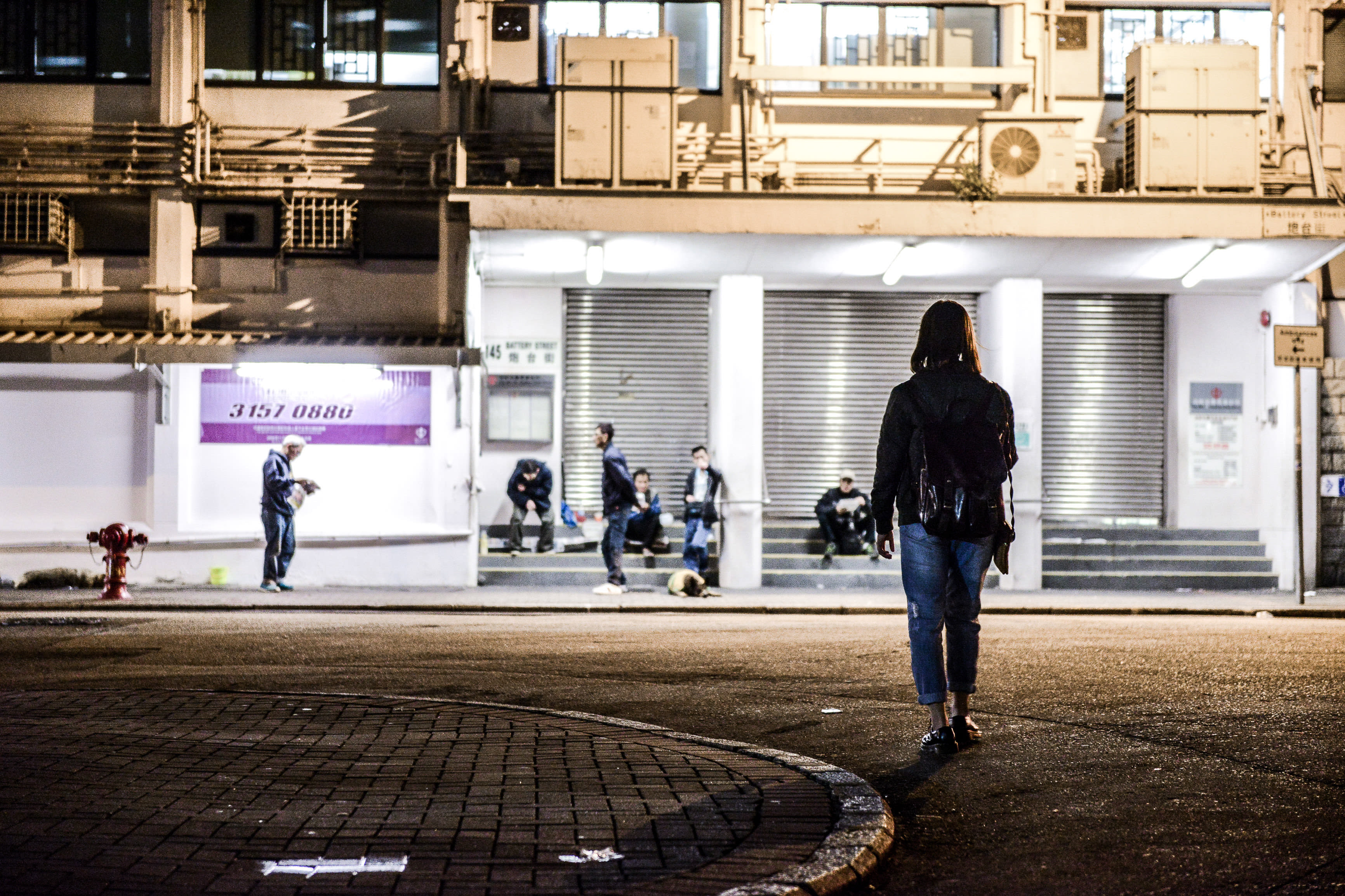 夜幕下的香港城市街頭，鏡頭前景中一人背對鏡頭，正走向一處燈火通明的建築入口，幾位人物正坐在階梯上休息。這種帶有工業風格的背景與靜謐、孤獨的氛圍，呼應了天台製作（Rooftop Productions）當代劇場作品《消失的海岸線》（Lost Shoreline）中引人深思的情感基調。《消失的海岸線》