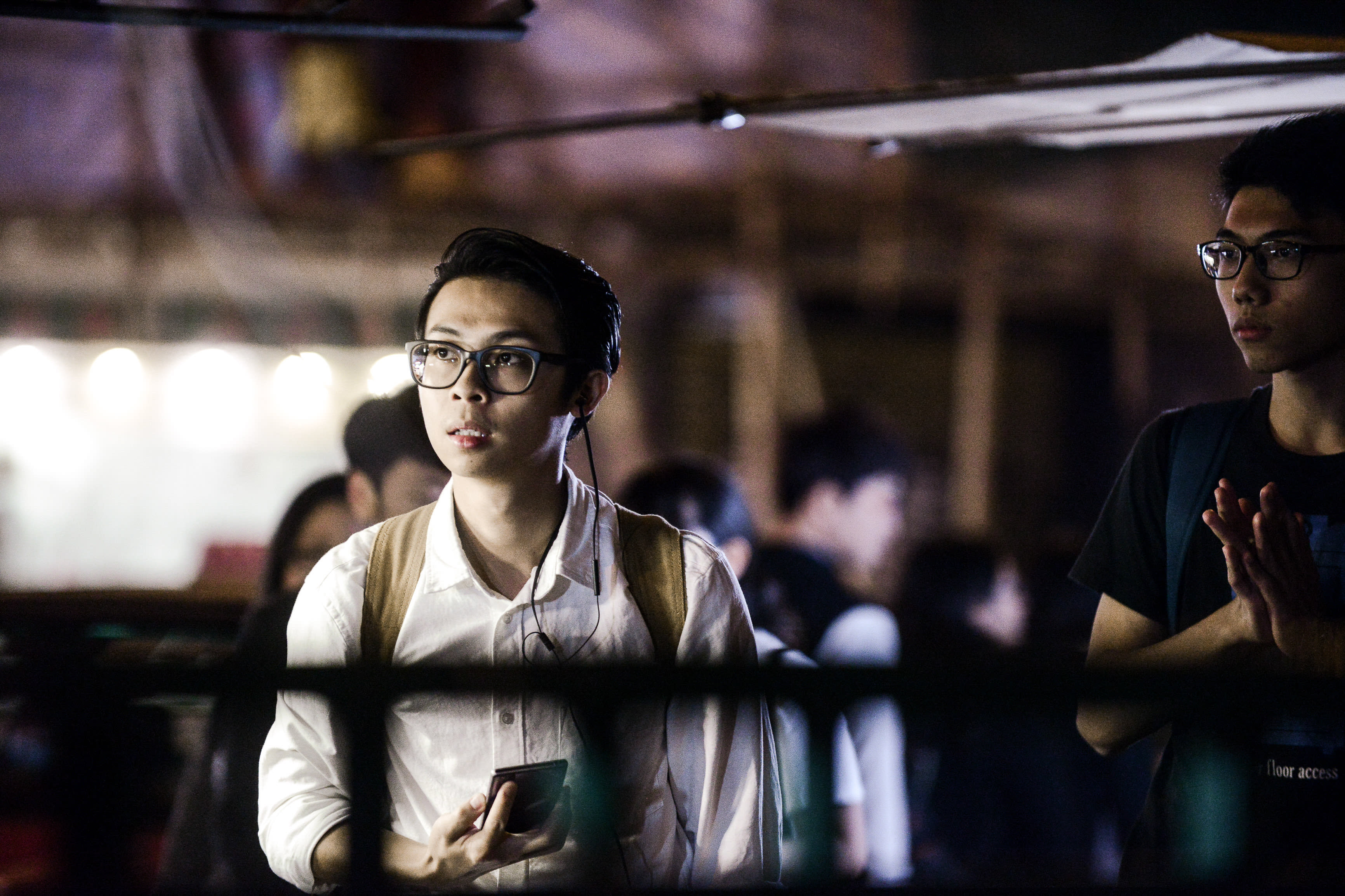 天台製作（Rooftop Productions）當代劇場作品《消失的海岸線》（Lost Shoreline）的排練劇照。演員趙展禧身穿簡潔的白襯衫並配戴眼鏡，手持手機並戴著耳機，神情專注沉思。舞台燈光營造出充滿戲劇感的氛圍，聚焦於他在幽暗背景下的表演瞬間。《消失的海岸線》