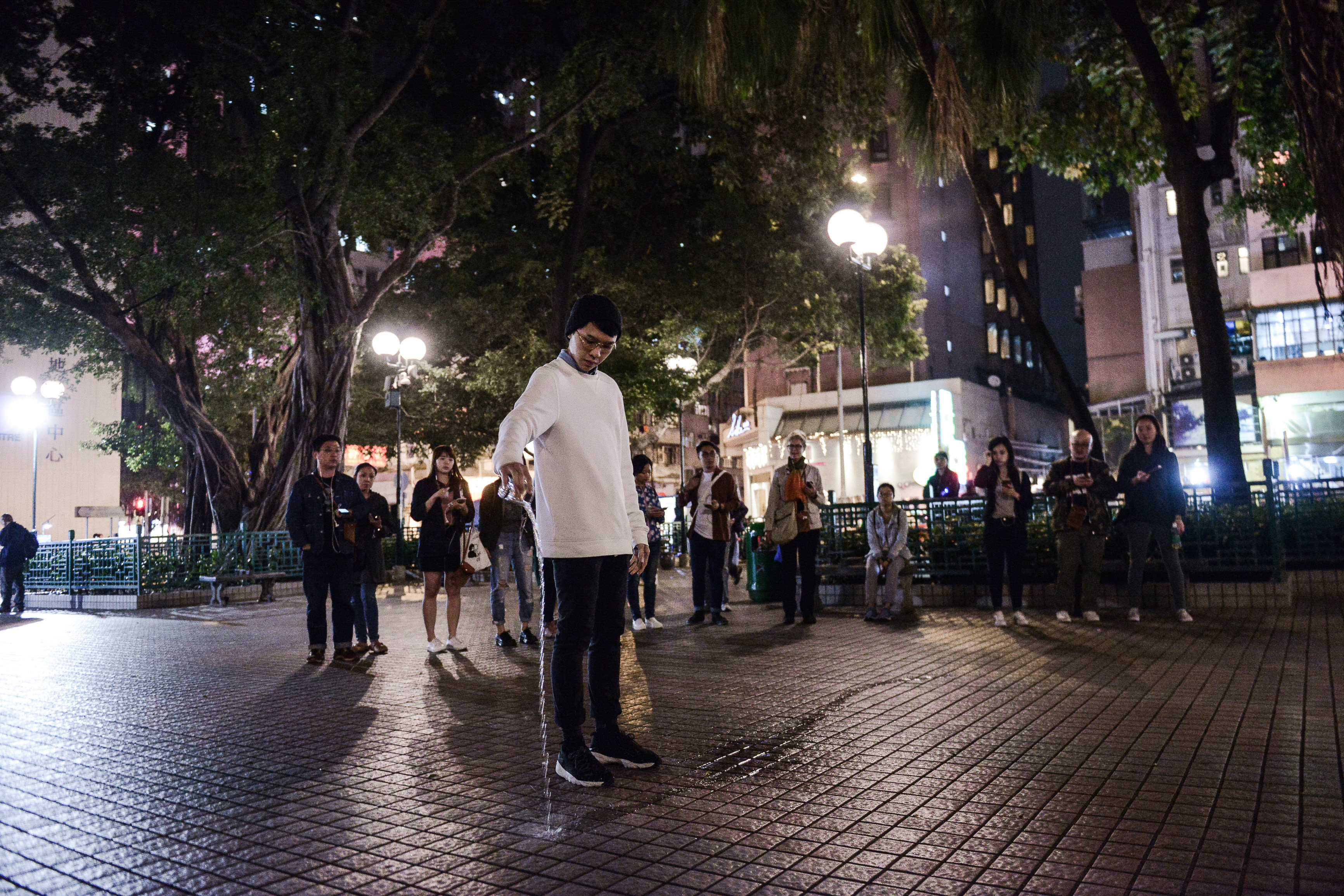 在香港的一個戶外公園，夜色昏暗，演員吳景隆身穿白色長袖上衣，正專注地將水從容器中緩緩倒在鋪滿地磚的地面上，這是天台製作當代劇場作品《消失的海岸線》中極具張力的一幕。周圍站著一圈靜默觀察的表演者，背景是茂密的樹木與城市燈火，深刻呼應了天台製作在城市空間中探討記憶與地景變遷的創作核心。「消失的海岸線」