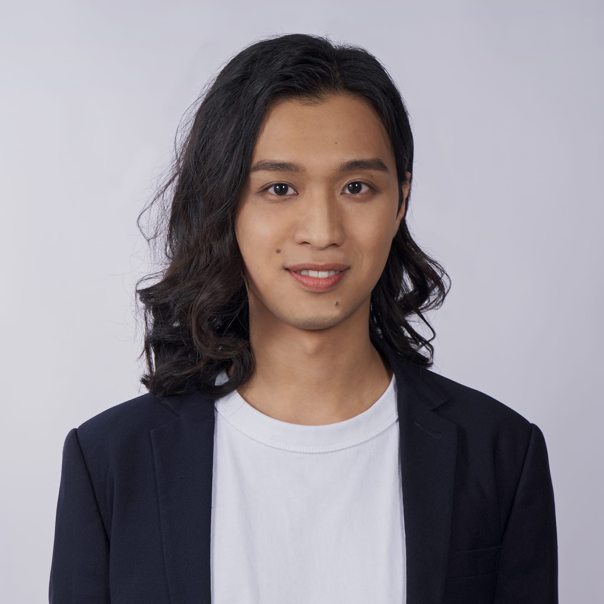 這是一張香港舞台劇演員麥晧鈿的專業形象照，背景為素雅的灰調攝影棚背景。照片中的他留著中長深色波浪捲髮，身穿白色圓領T恤外搭深色西裝外套，正對鏡頭展露親切的微笑。這張照片展現了這位演員的個人魅力，他也是天台製作（Rooftop Productions）當代香港劇場演出中常見的表演者。