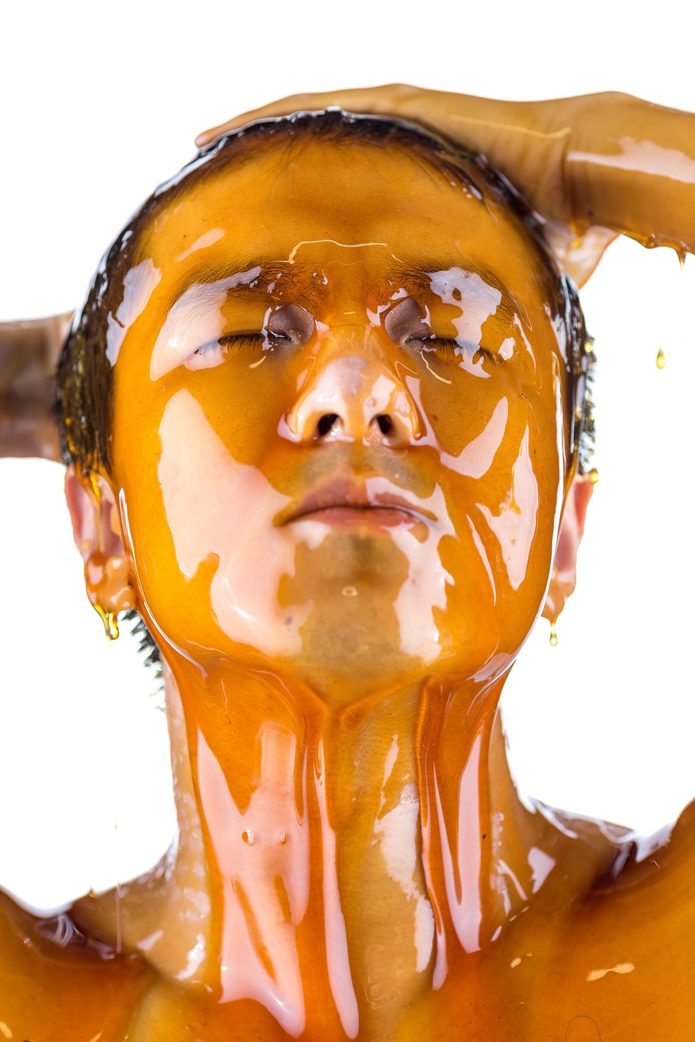 A dramatic close-up promotional photograph for the contemporary Hong Kong theatre production 'Milk and Honey'. Actor Billy Sy is captured from the chest up, his head tilted back with his eyes closed in a moment of intense, raw emotion. Thick, amber-colored honey cascades over his face, hair, and neck, catching the studio light and creating a stark, high-contrast effect against a clean white background. This evocative imagery serves as the promotional photo of Billy Sy for Milk and Honey.