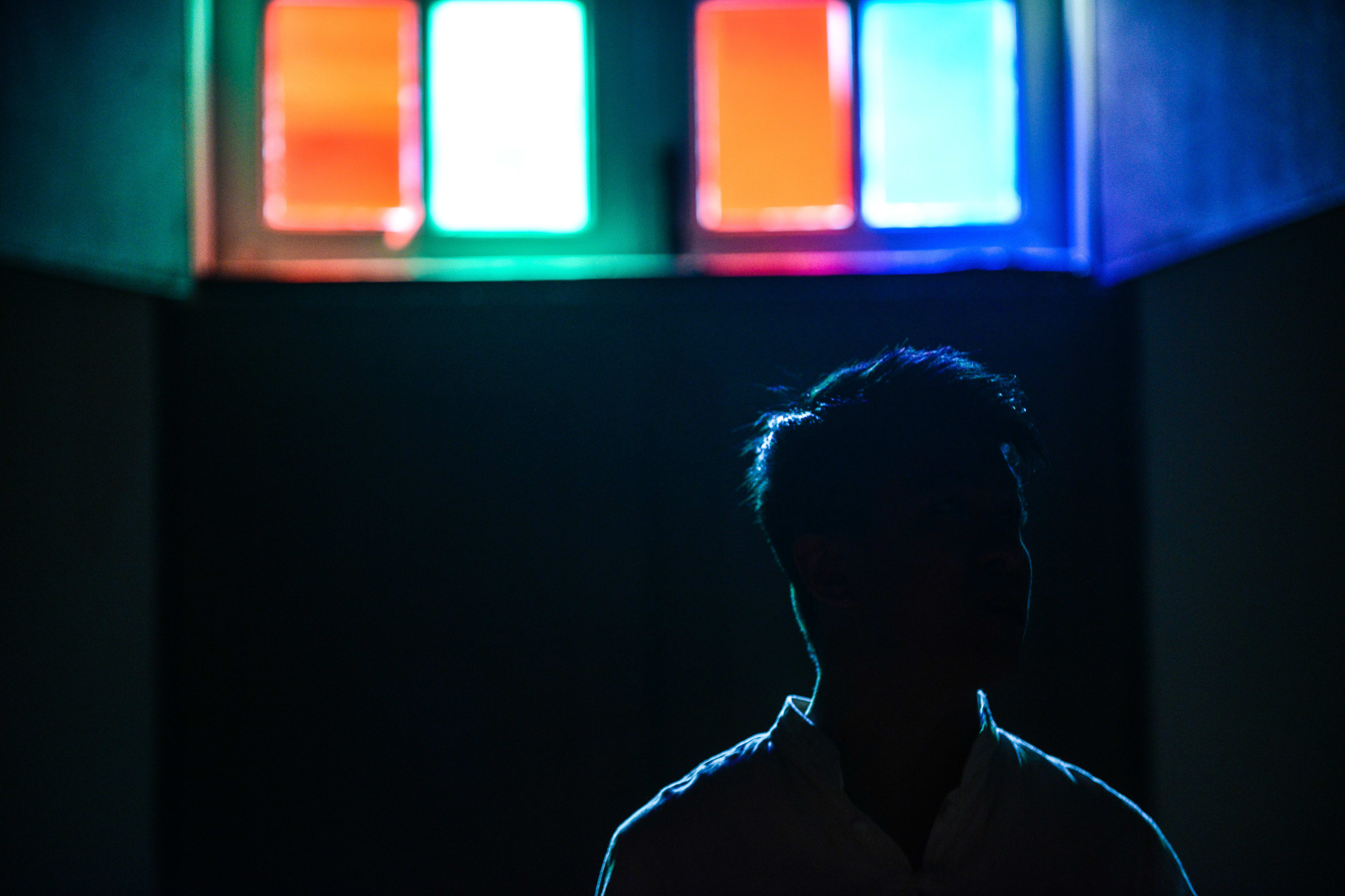 A dramatic, moody portrait of actor Billy Sy (施標信) in a scene from the Rooftop Productions (天台製作) contemporary theatre piece, Milk and Honey (《牛奶與蜜糖》). Sy is captured in silhouette against a stark, dark background, his form outlined by a sharp blue rim light. Above him, four glowing rectangular panels cast vibrant, saturated hues of orange, white, and blue, creating a striking contrast that emphasizes the theatrical atmosphere. Milk and Honey.