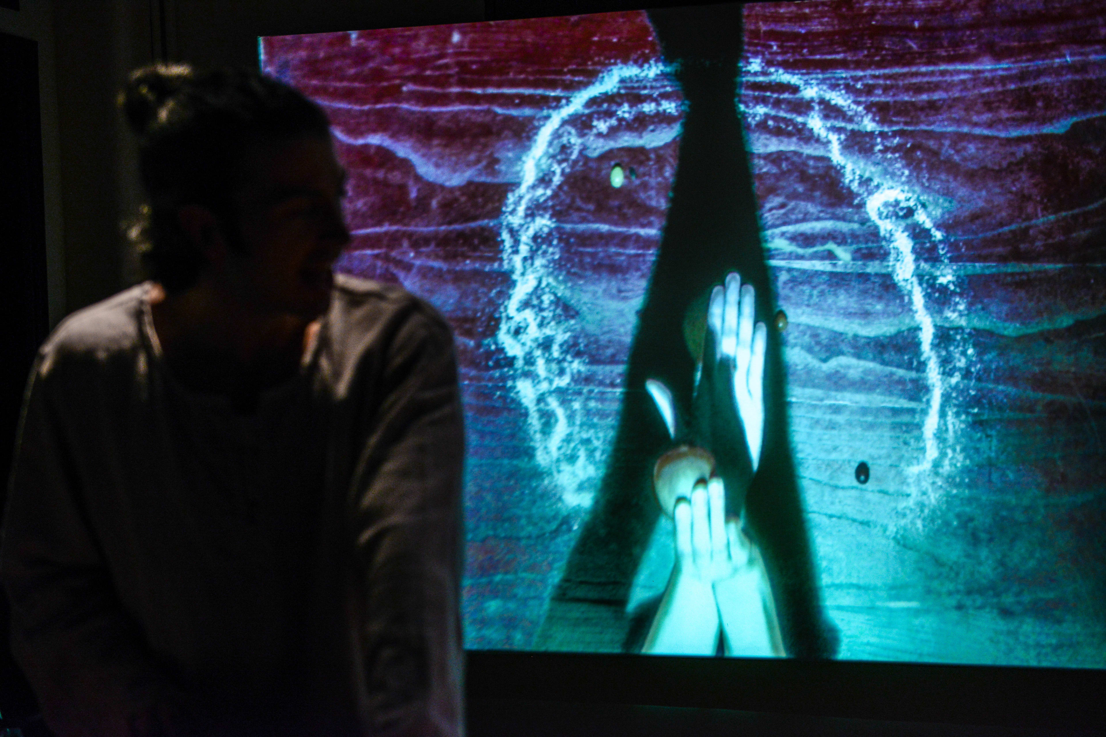 In a dimly lit stage setting for Rooftop Productions' contemporary theatre piece 'Milk and Honey', performer Ivor Houlker is seen in profile. Behind him, a large screen projects a striking, ethereal visual of hands held together in a gesture of supplication or prayer, framed by a luminous, swirling ring of light that resembles a cosmic portal. The contrast between the shadows and the vibrant teal and deep red hues of the projection emphasizes the experimental nature of this Hong Kong stage performance. 'Milk and Honey'