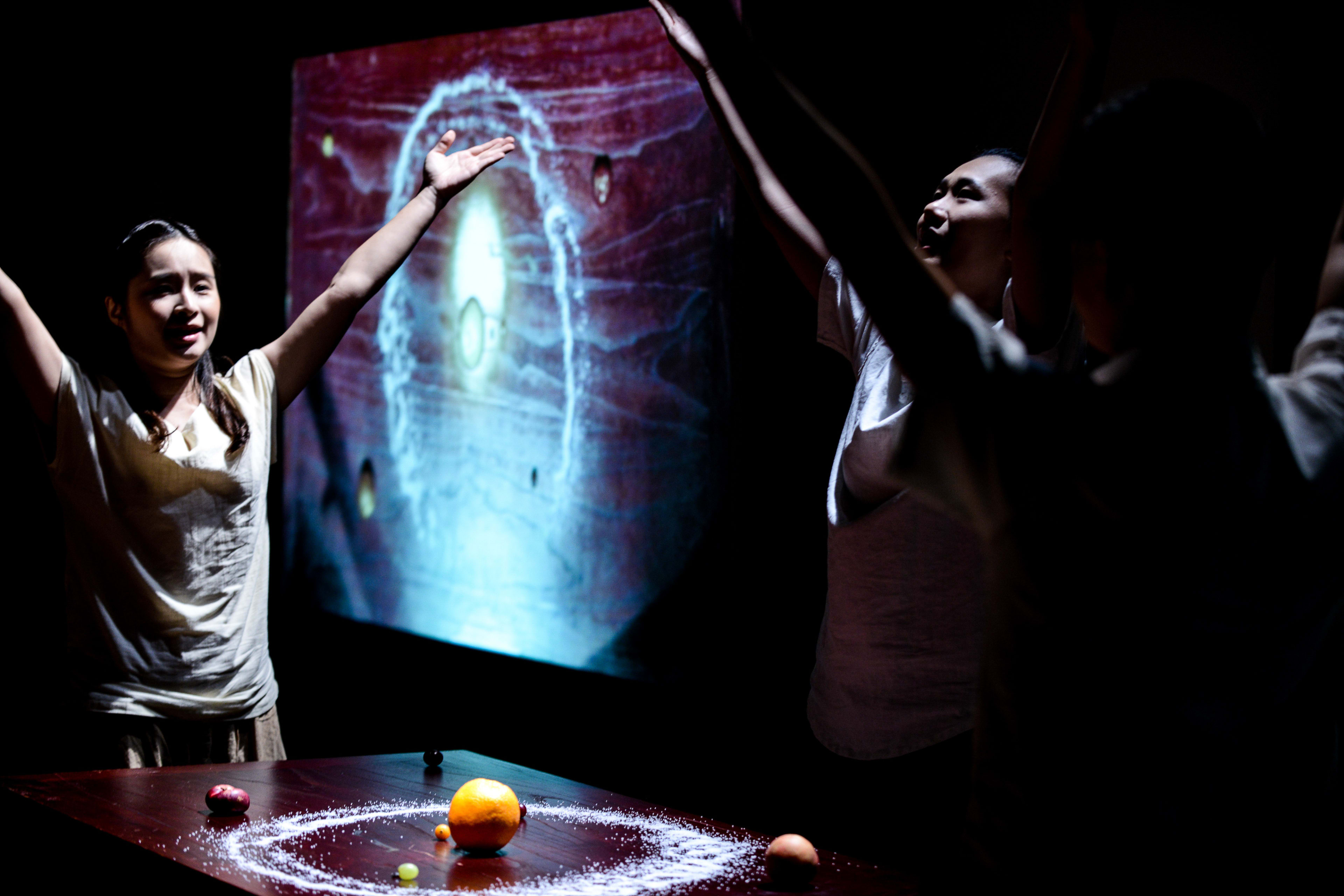 In a scene from the Rooftop Productions contemporary theatre piece Milk and Honey, actors Michelle Li and Lung Jes stand behind a table illuminated by a circular display of grains. The performers reach their arms upward in a dramatic gesture, mirroring the swirling, cosmic-like projection of a milk-and-honey aesthetic on the screen behind them. The lighting is moody and ethereal, highlighting the intimate and abstract nature of this Hong Kong stage production. "Milk and Honey"