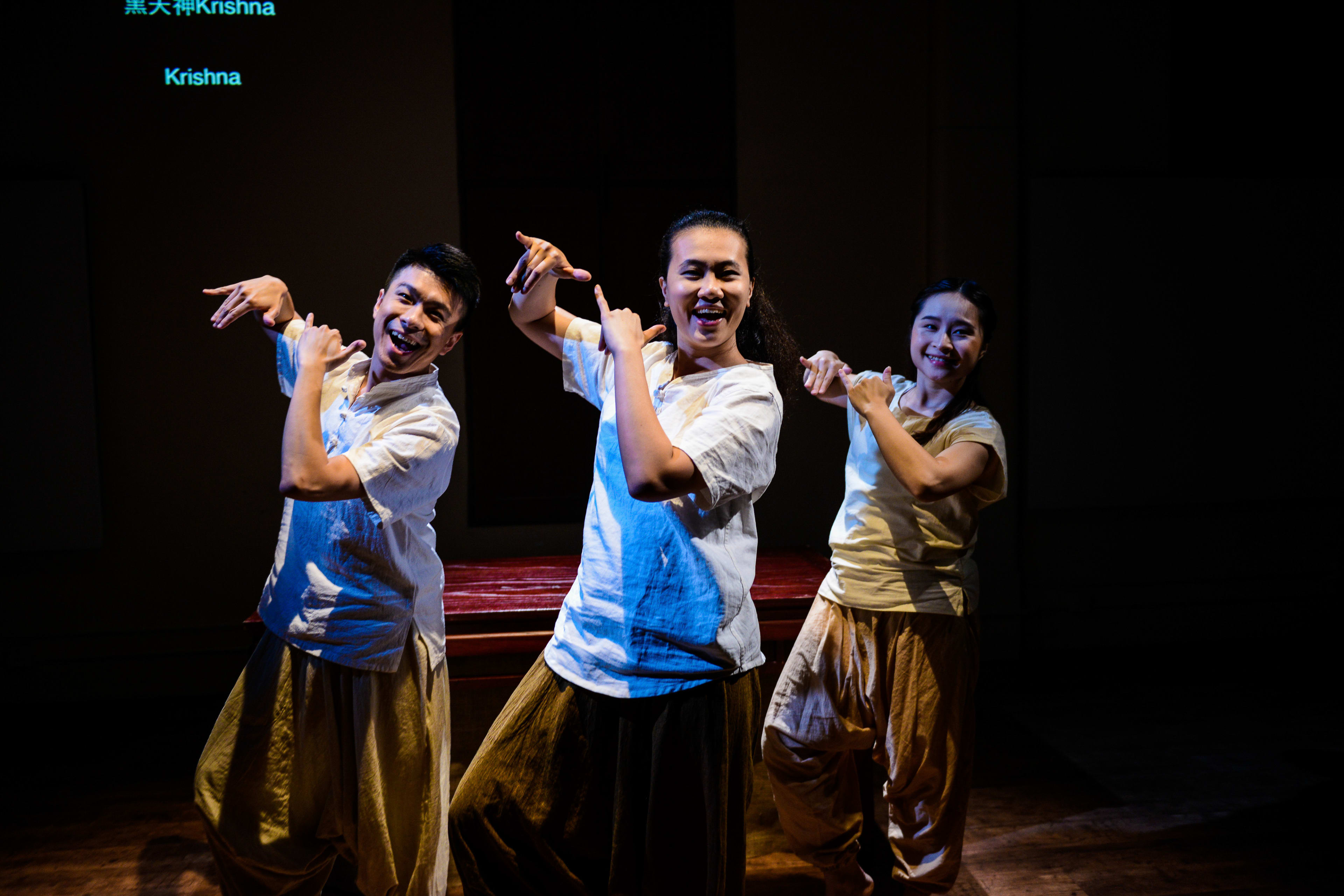 Three actors perform a choreographed dance sequence in Rooftop Productions' contemporary theatre piece 'Milk and Honey'. Dressed in matching cream-colored shirts and loose trousers, they strike expressive poses against a dark, minimalist stage background under moody spotlighting. The actors—Billy Sy, Michelle Li, and Lung Jes—capture a moment of thematic significance from the production. "Milk and Honey"