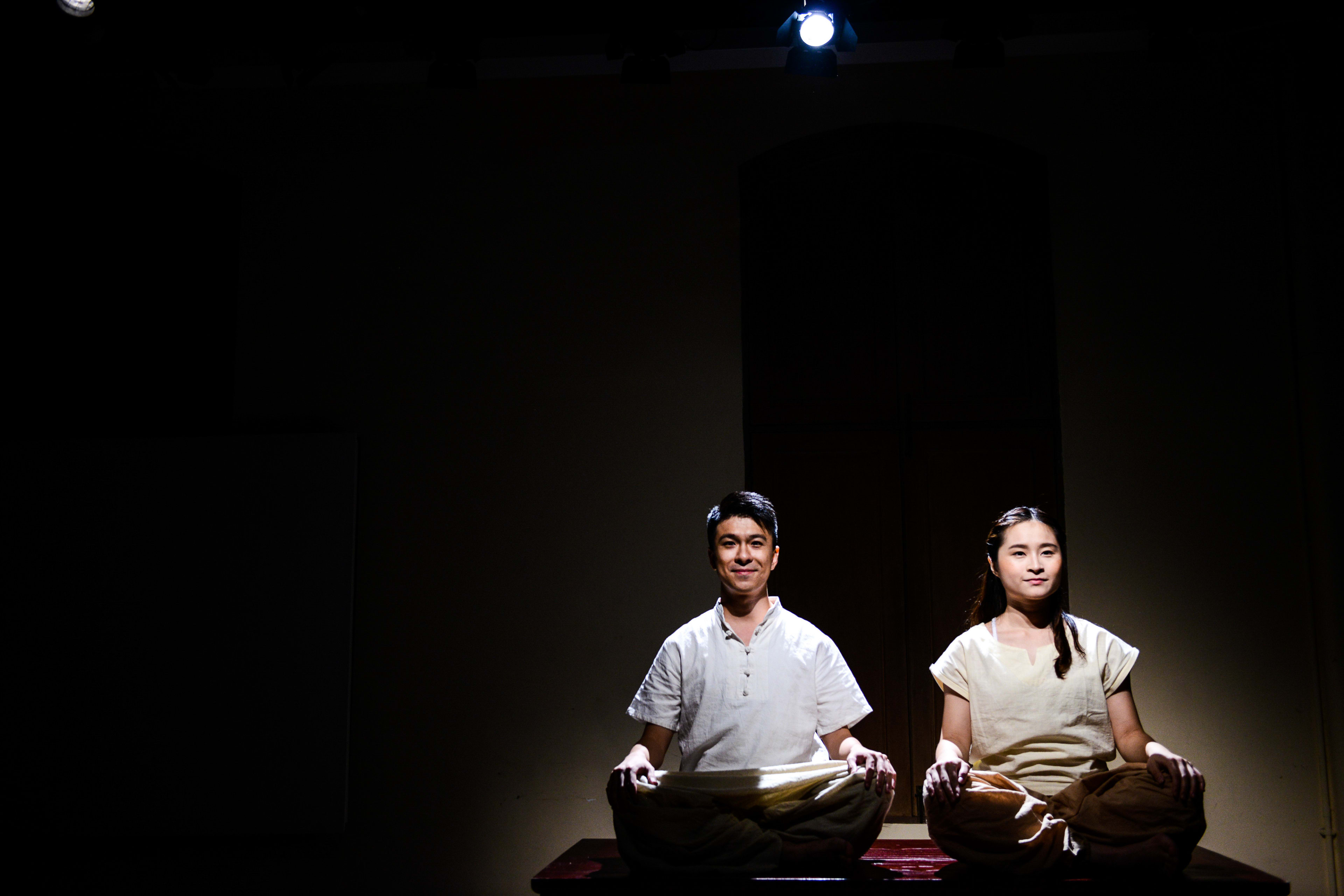 Actors Billy Sy and Lung Jes perform a meditative scene in Rooftop Productions' contemporary theatre piece, Milk and Honey. They sit cross-legged on a raised platform, illuminated by a stark, singular spotlight against a dark background, embodying the minimalist staging characteristic of Hong Kong's experimental stage. "Milk and Honey"