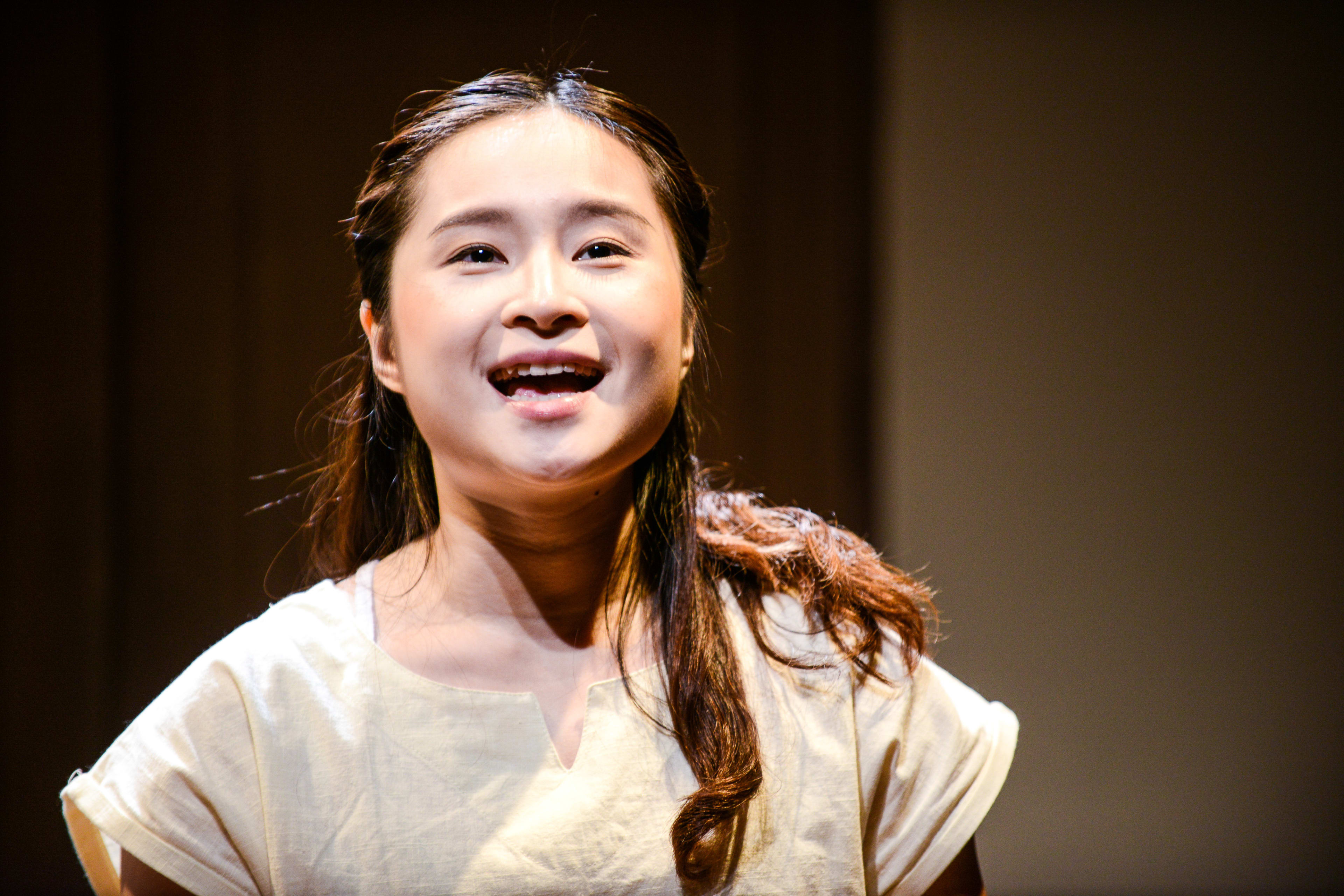 Close-up of actor Lung Jes performing on stage during Rooftop Productions' contemporary theatre piece, Milk and Honey. She is lit by a warm spotlight, wearing a simple cream-colored top, with an emotive, joyful expression as she sings or speaks during the play. "Milk and Honey"