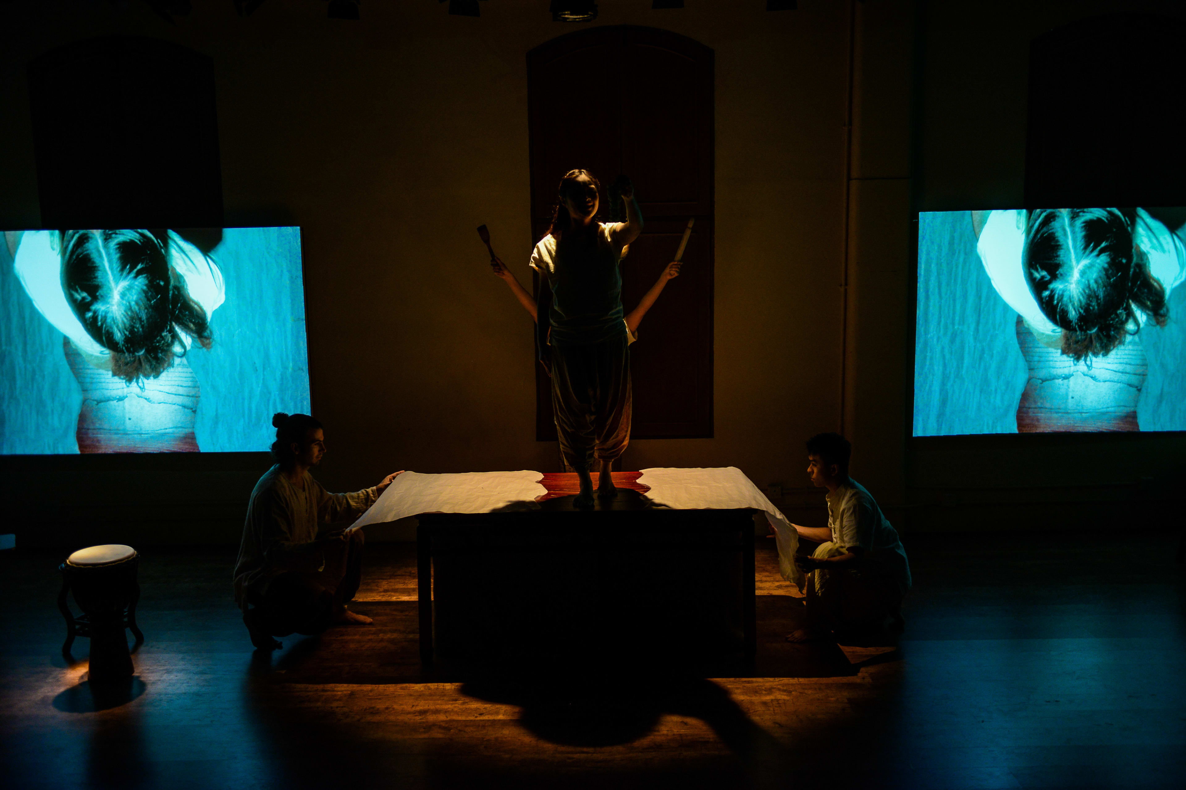在這張天台製作（Rooftop Productions）當代劇場作品《牛奶與蜜糖》（Milk and Honey）的劇照中，演員隆藹宜站在中央的桌子上，身姿在溫暖且極具戲劇性的聚光燈下形成剪影。另外兩名表演者分別蹲在桌子的兩端，雙手按在桌面上，彷彿在為空間提供支撐。舞台兩側的大型屏幕投射出單色且抽象的人體影像，展現了香港獨立劇場簡約而震撼的視覺美學。《牛奶與蜜糖》