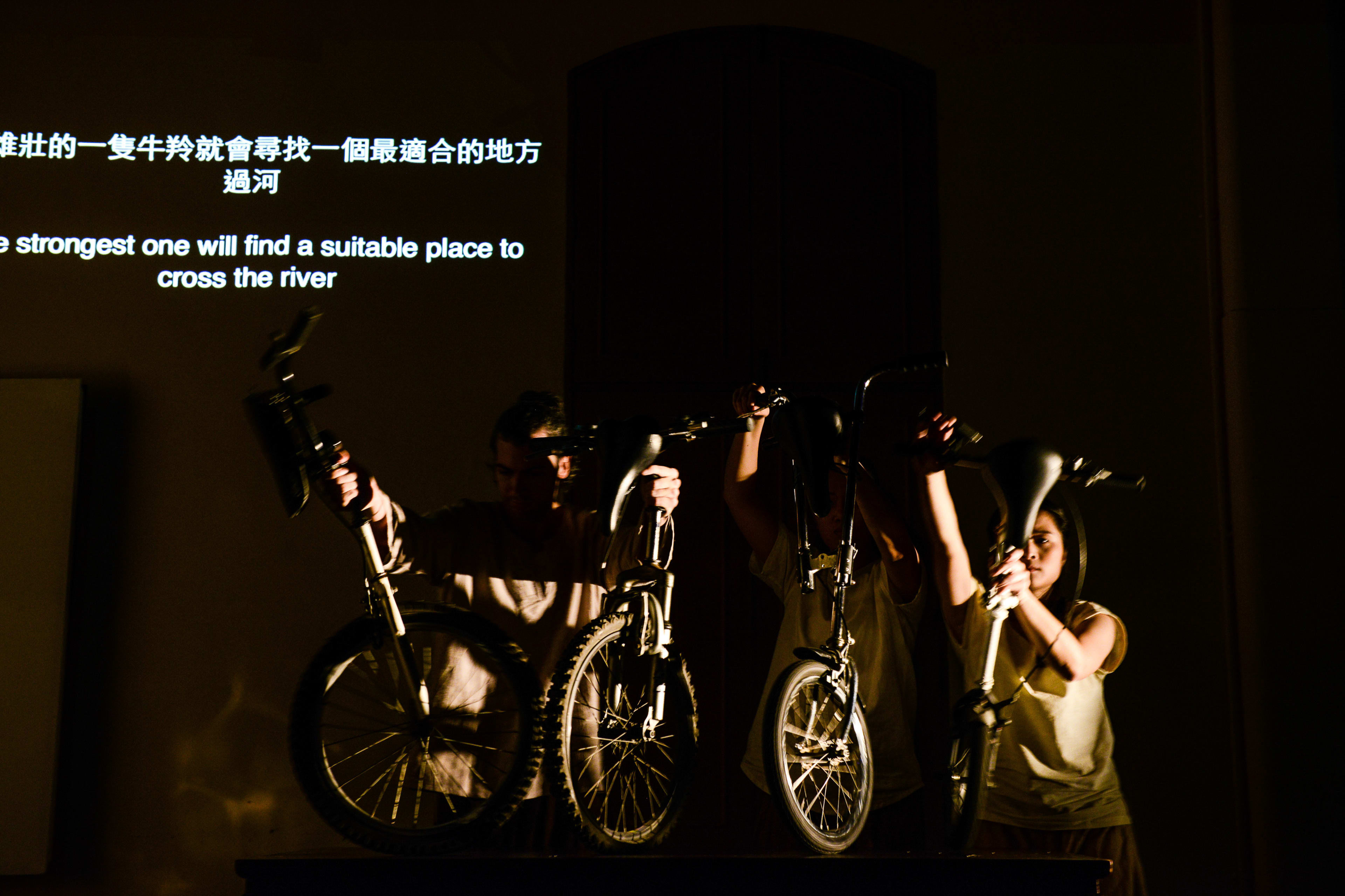 In this evocative scene from the Rooftop Productions (天台製作) contemporary theatre piece 'Milk and Honey', two actors are silhouetted against a dark stage, each holding a bicycle aloft to signify a symbolic journey. The dramatic low-key lighting highlights their forms and the metallic frames of the bikes, creating a striking visual metaphor for migration and resilience. Behind them, a projected subtitle reads: 'The strongest one will find a suitable place to cross the river.'