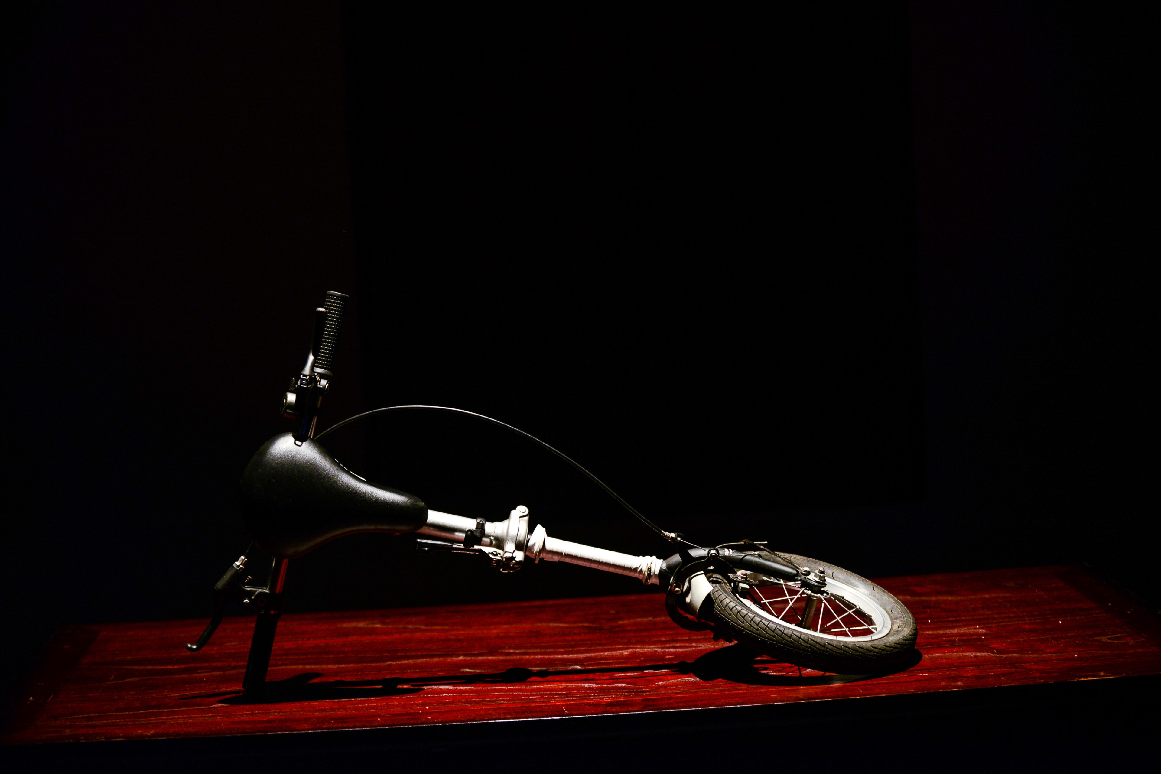 A minimalist, high-contrast promotional image for the Rooftop Productions contemporary theatre play 'Milk and Honey'. A single folded bicycle, representing a prop from the stage performance, rests on a dark wooden surface against a stark black background, illuminated by a single spotlight that emphasizes the loneliness and fragility of the object. 'Milk and Honey'.