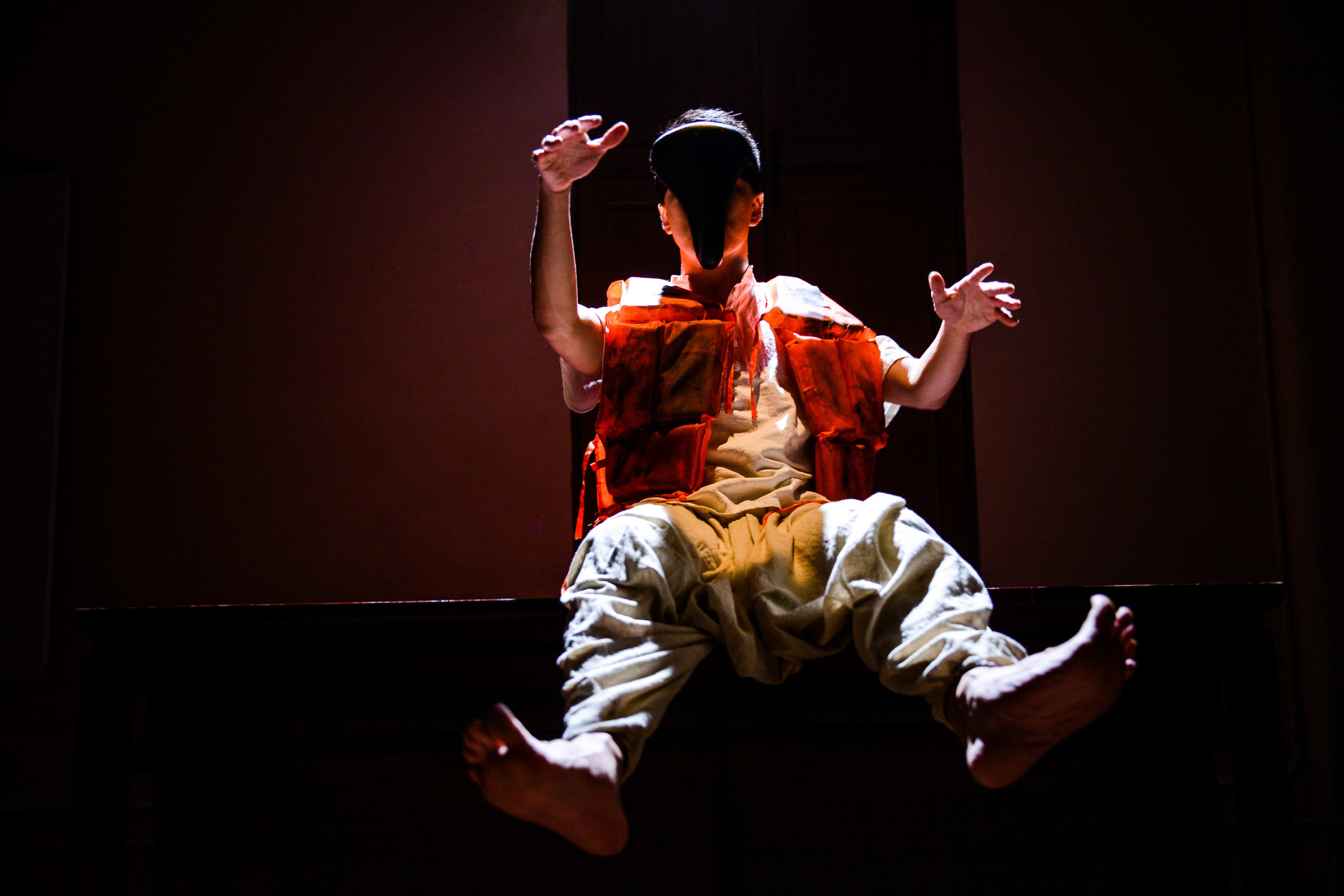 A performer, Billy Sy, sits suspended in mid-air against a dark background, illuminated by a sharp spotlight in this dramatic scene from the Rooftop Productions contemporary theatre play, Milk and Honey. He is wearing a bright orange life vest over light-colored clothing and a mysterious black mask that obscures his face, evoking a surreal and unsettling mood. The lighting highlights his bare feet and outstretched hands as he floats, capturing a powerful moment of physical theatre.