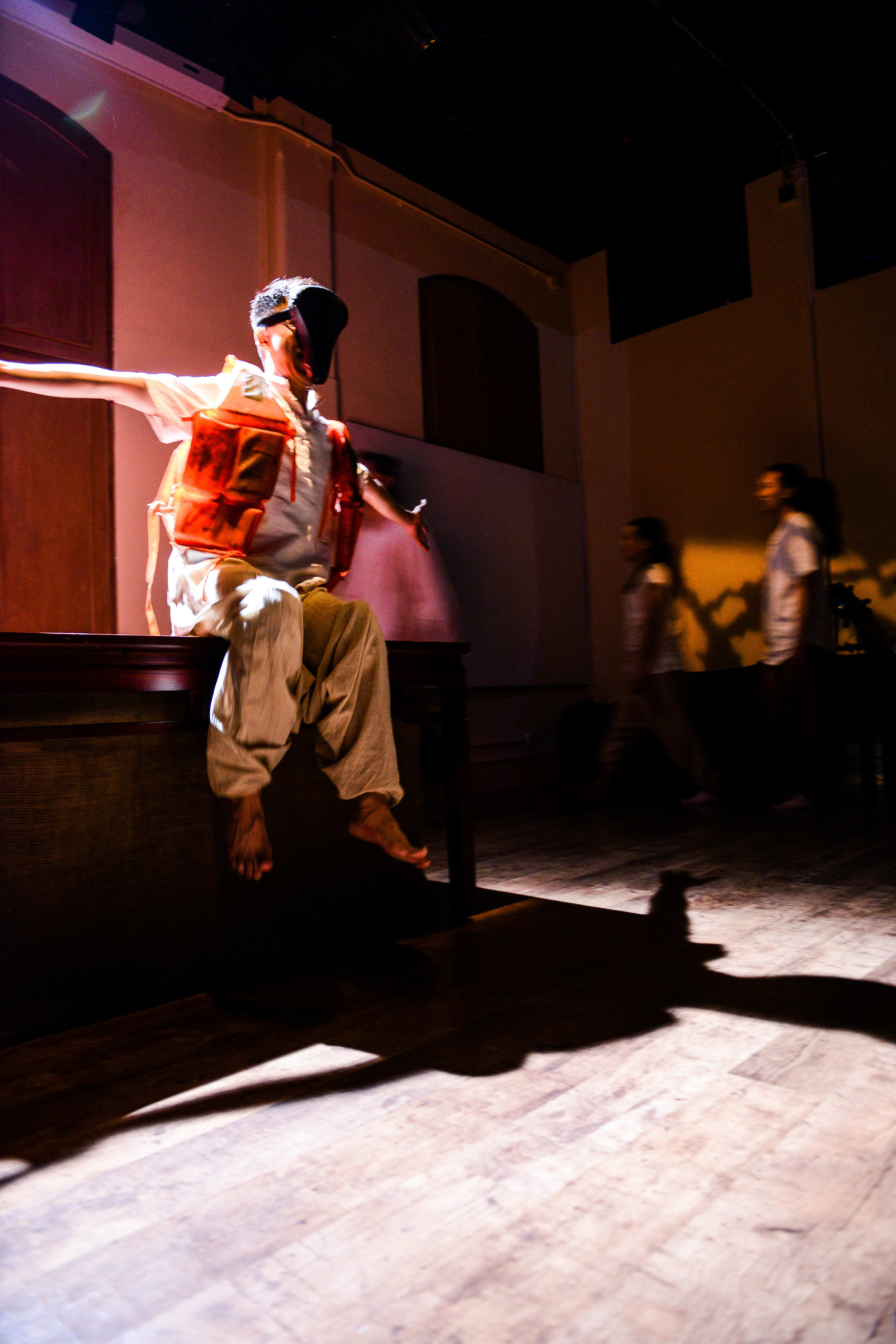 A dramatic stage photo from Rooftop Productions' contemporary theatre piece, Milk and Honey (《牛奶與蜜糖》). In a dimly lit, moody performance space, actor Billy Sy sits perched on the edge of a desk, wearing a bright orange life vest and an abstract black face mask, his arms outstretched in a gesture of vulnerability or surrender. In the darkened background, two other performers move across the stage, their blurred figures adding to the tension of the scene. Milk and Honey.