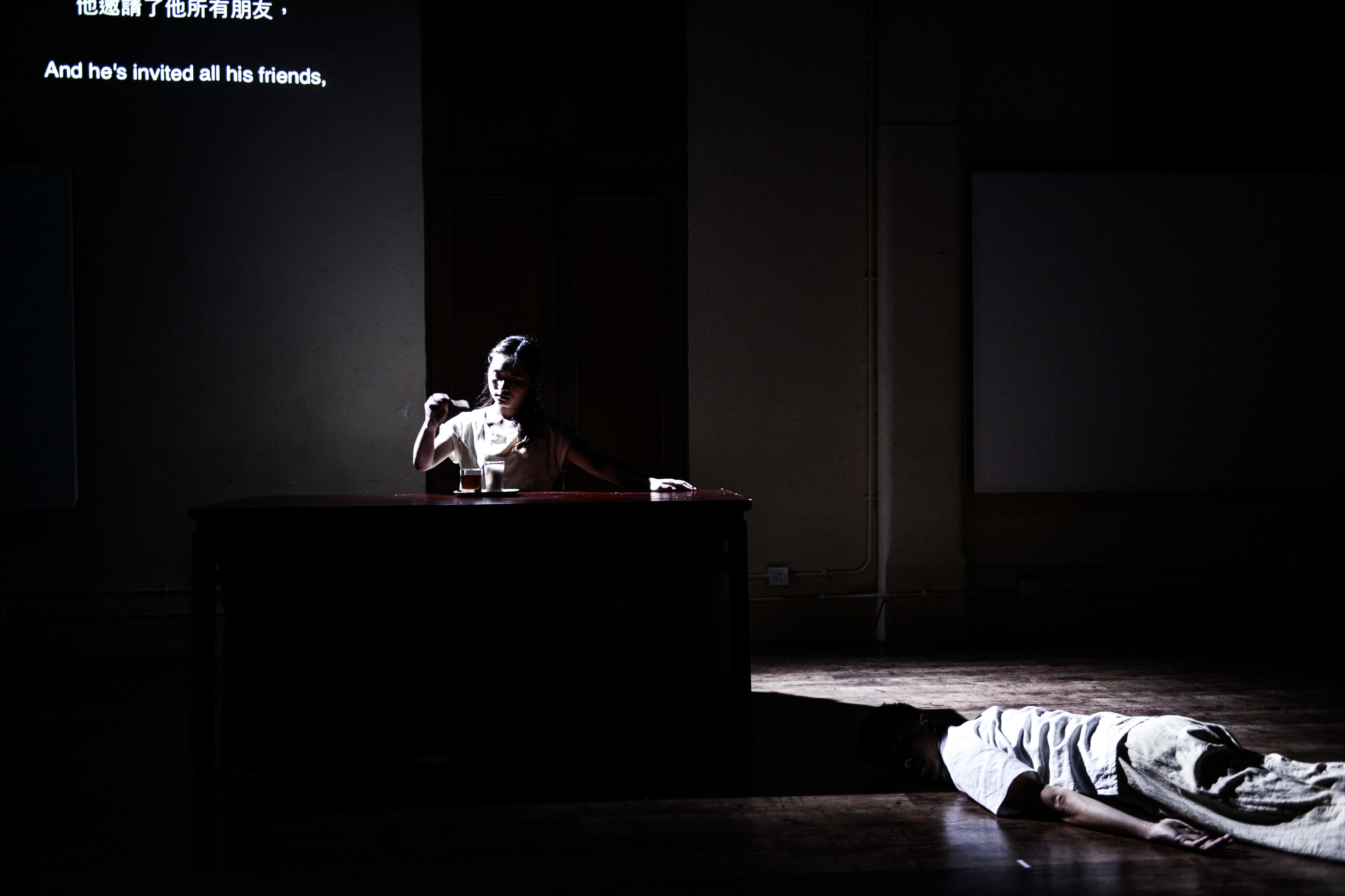 在天台製作的當代劇場作品《牛奶與蜜糖》（Milk and Honey）中，一名演員在黑暗舞台中央的聚光燈下端坐在書桌旁，手中拿著物品，神情專注；舞台前方則有另一名演員躺在地板上，形成強烈的視覺對比與戲劇張力。台上演員低語：「他邀請了他所有朋友。」