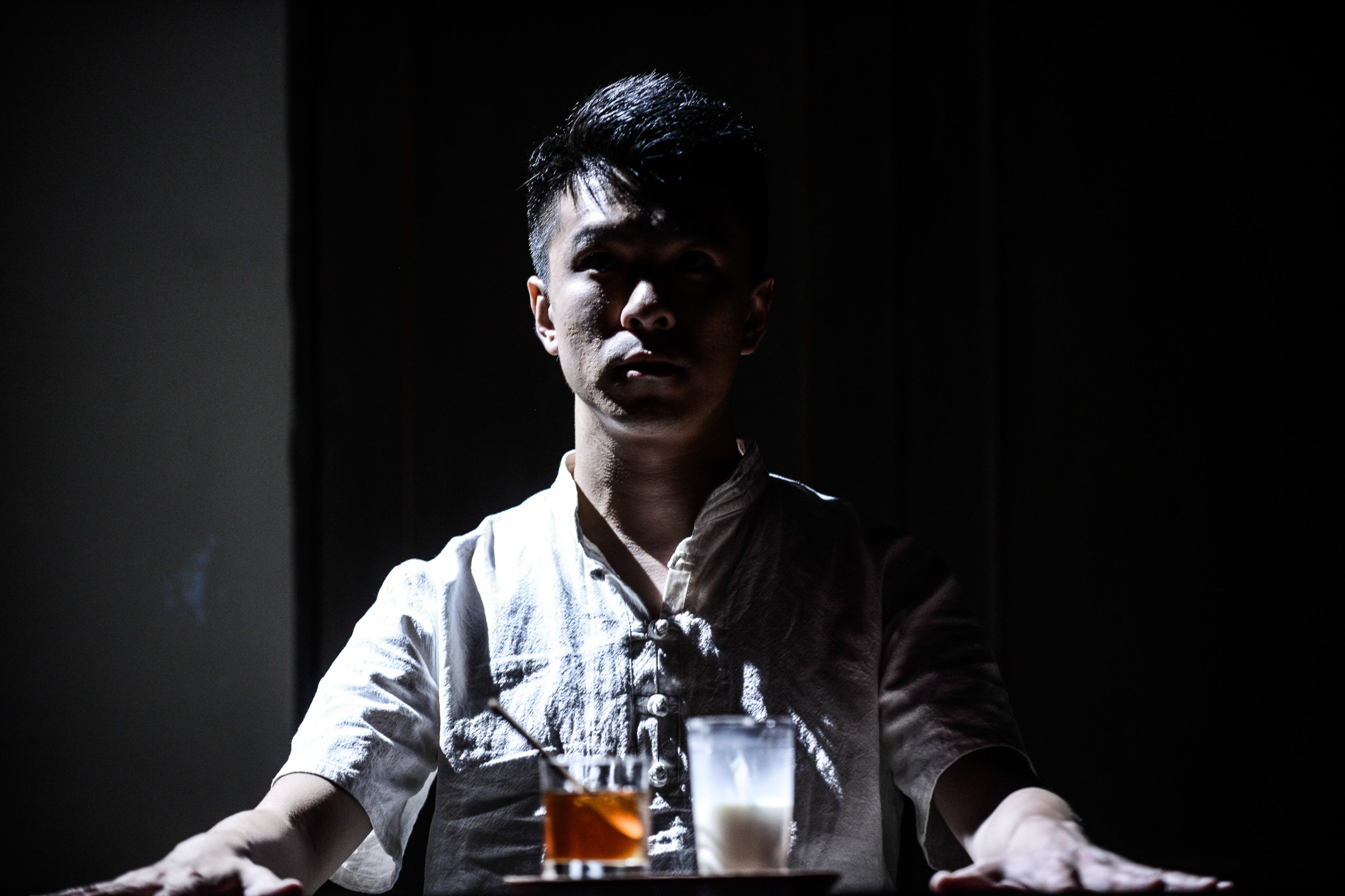 In this dramatic promotional shot for Rooftop Productions' contemporary theatre piece 'Milk and Honey', actor Billy Sy is illuminated by a sharp, high-contrast spotlight against a pitch-black background. He sits at a table with his hands outstretched, appearing solemn and contemplative, with a glass of amber liquid and a glass of milk placed before him. This atmospheric image captures the tense, introspective mood of the show. 'Milk and Honey'