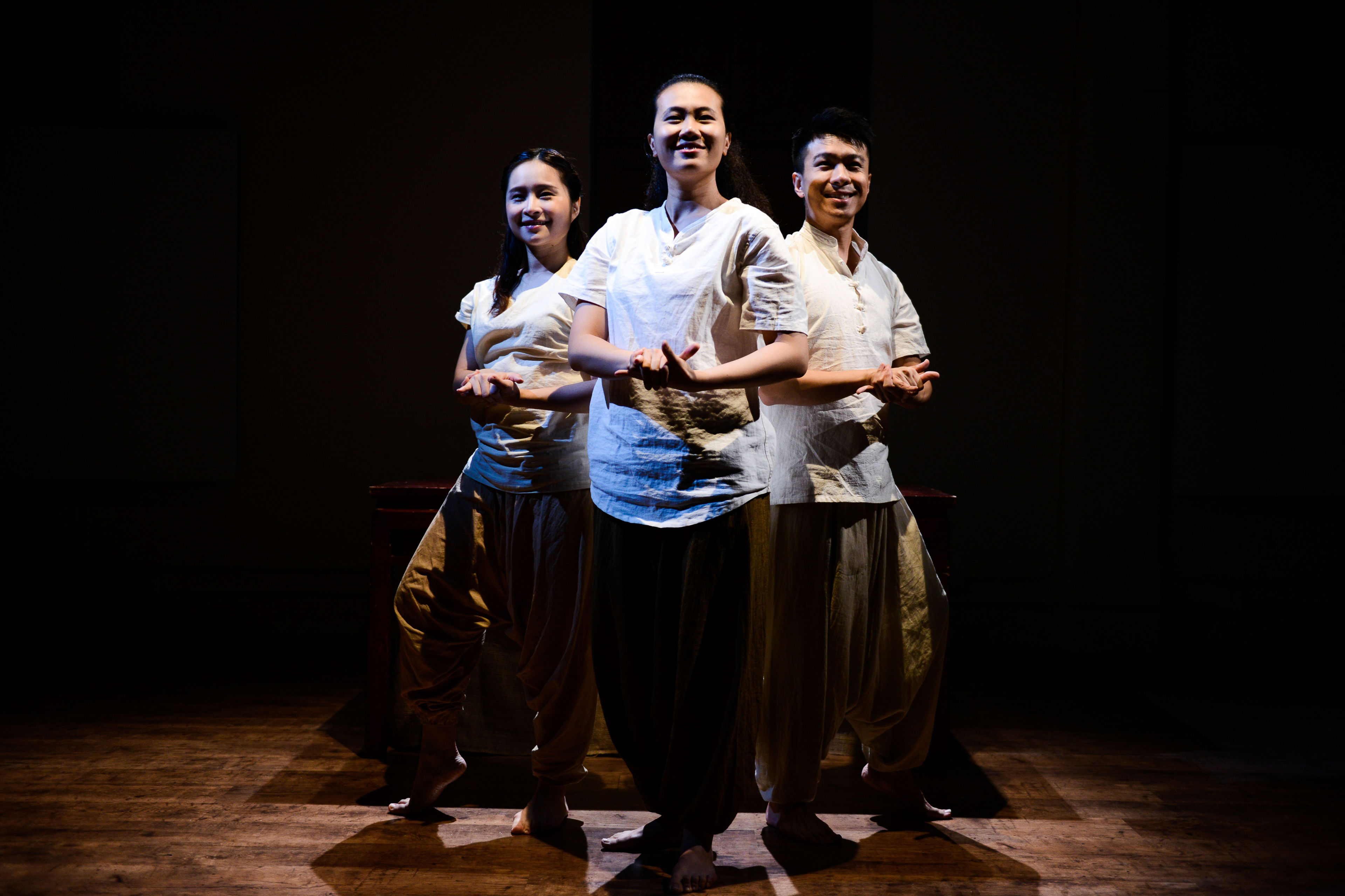天台製作（Rooftop Productions）的三位演員站在昏暗、簡約的木製舞台上，在當代香港劇場作品《牛奶與蜜糖》的排練中，身處聚光燈下。演員們身穿簡約的白色襯衫與寬鬆長褲，神情專注而平和，展現出協調一致的肢體動作。居中的女演員雙手在胸前優雅交疊，另外兩位演員則稍稍靠後，構成了一幅平衡且富有感染力的舞台構圖。《牛奶與蜜糖》