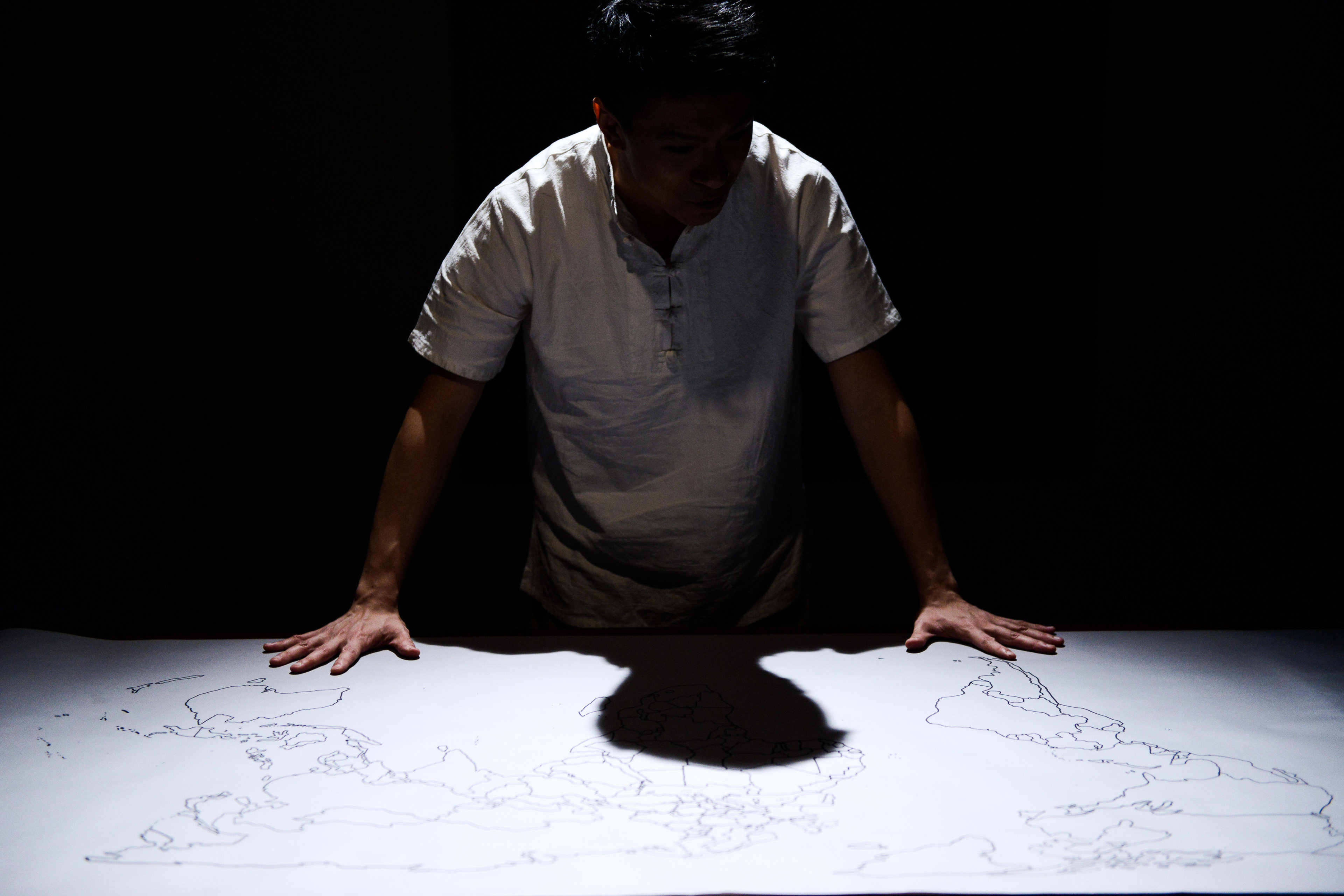 天台製作（Rooftop Productions）當代劇場作品《牛奶與蜜糖》（Milk and Honey）的宣傳劇照。舞台上燈光昏暗，一位男性表演者俯身站在桌前，雙手按在鋪滿白紙的地圖輪廓上；頭頂的強光將他的身影投射在畫面上，營造出強烈的視覺對比與孤獨感，展現出關於遷徙與連結的劇場張力。《牛奶與蜜糖》
