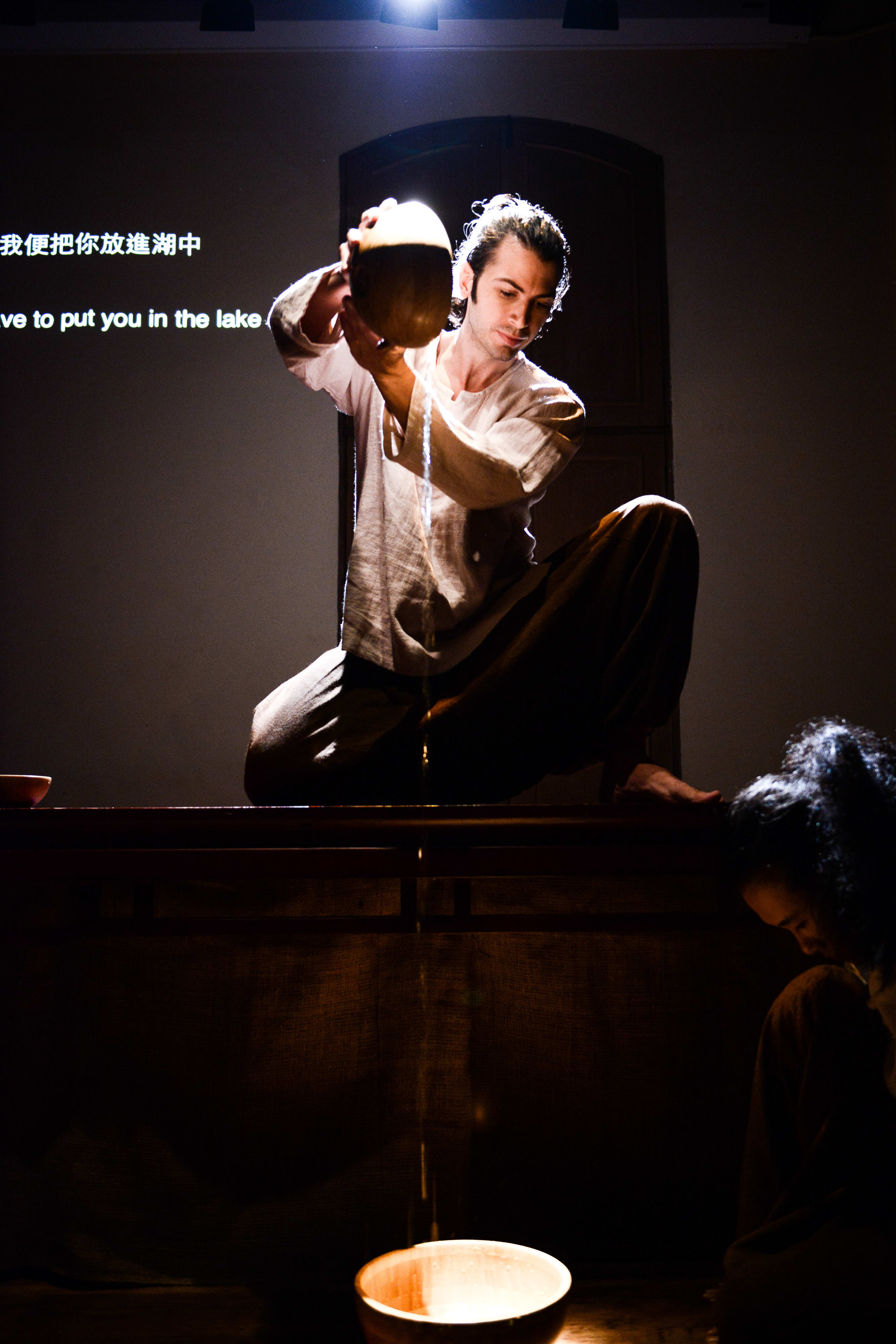 在這場由天台製作（Rooftop Productions）呈獻的當代劇場作品《牛奶與蜜糖》中，演員艾浩家（Ivor Houlker）蹲在木製平台上，背後映襯著充滿戲劇感的強烈聚光燈。他正小心翼翼地將液體從一個質樸的木製器皿倒入下方的碗中，形成一道精緻的光束。旁邊的陰影中隱約可見另一位表演者。舞台背景投影著雙語字幕，其中一句寫道：「我便把你放進湖中」。表演者唸道：「我便把你放進湖中」。