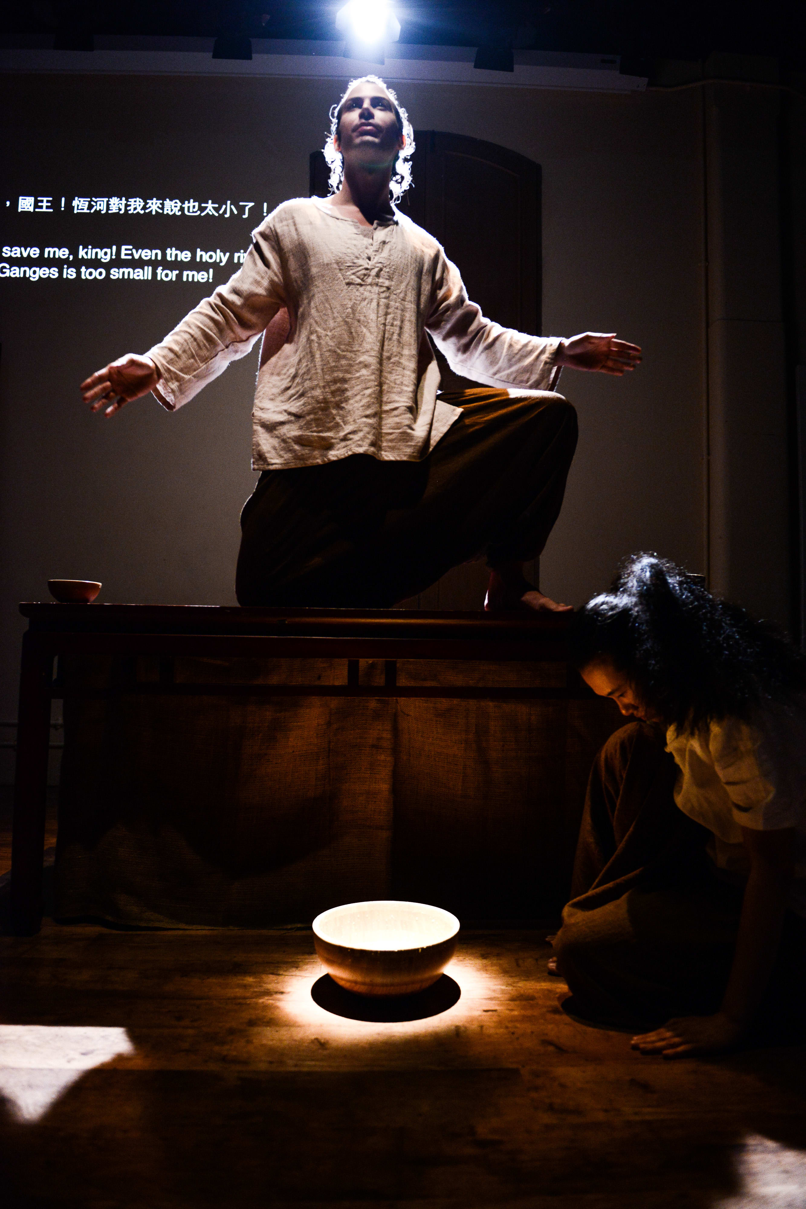 在天台製作的當代劇場作品《牛奶與蜜糖》中，演員艾浩家（Ivor Houlker）站在木桌上，雙臂向外延伸，呈現出一種神聖激昂的姿態。在他下方，另一位表演者在舞台地板上一盞發光的缽旁跪地沉思。背景投影上的字幕寫著：「國王！恆河對我來說也太小了！」