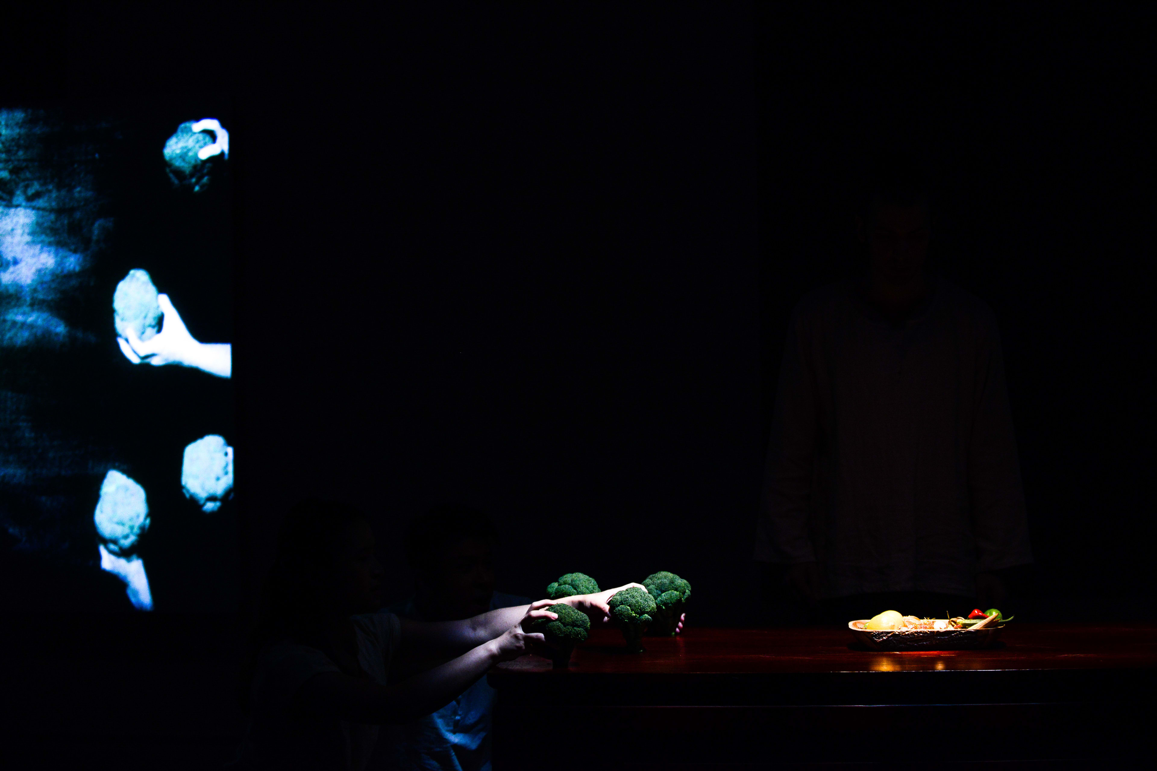 In this atmospheric scene from Rooftop Productions' contemporary theatre piece 'Milk and Honey', a performer sits in near-total darkness at a wooden table, illuminated by a warm spotlight. The performer's hands are visible as they interact with clusters of broccoli, while a digital projection on the left wall displays ghostly, repeated images of hands holding similar round objects. The low-key lighting and minimalist staging highlight the company's signature experimental style. 'Milk and Honey'