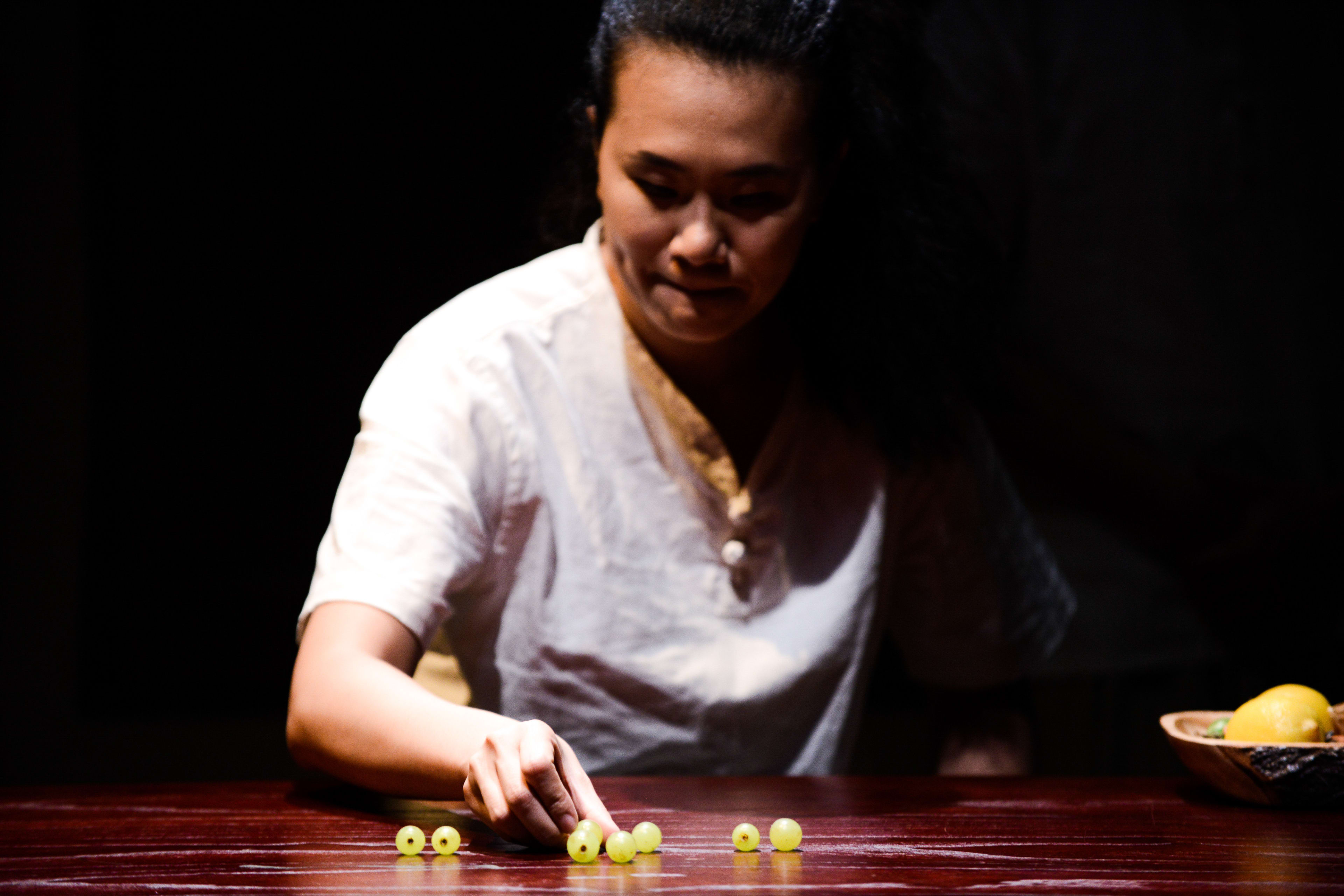 在這張天台製作（Rooftop Productions）當代劇場作品《牛奶與蜜糖》的劇照中，演員李婉晶坐在一張深色光亮的木桌前，在漆黑的背景下，一束聚光燈聚焦在她身上。她全神貫注地凝視並輕觸著桌上一小堆排列過的青葡萄，展現出親密而細膩的戲劇張力，突顯了該劇極簡主義的舞台美學。當她若有所思地望著葡萄時，輕聲道出：「牛奶與蜜糖」。