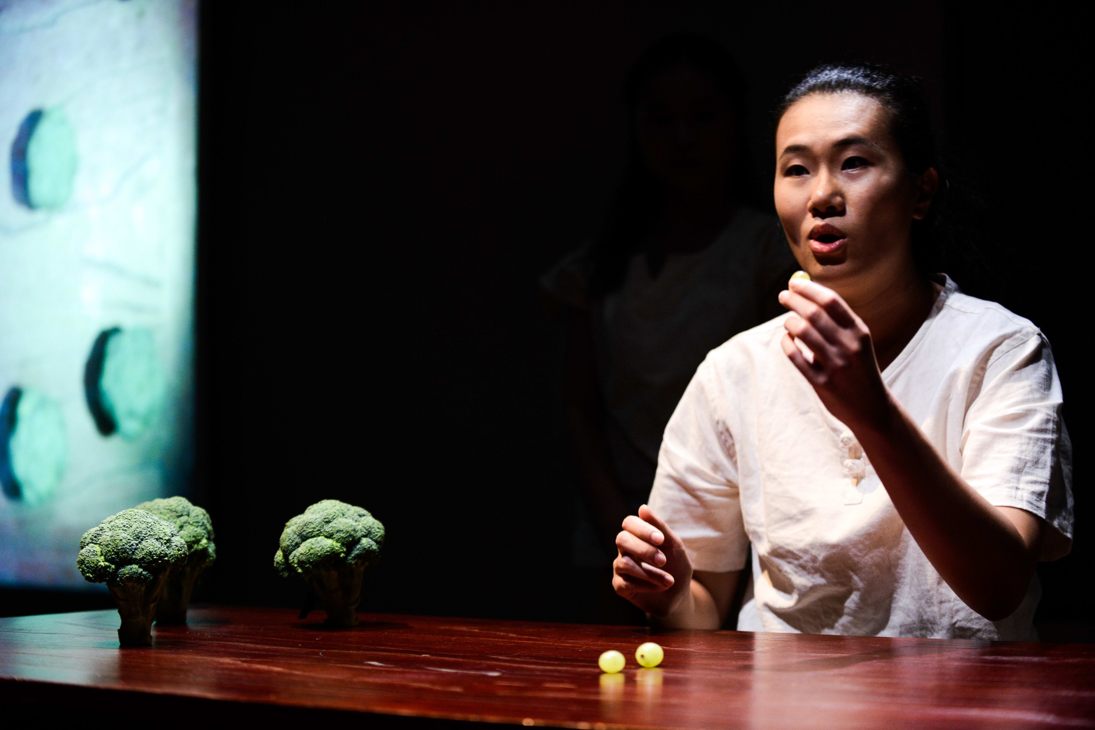 在天台製作的當代劇場作品《牛奶與蜜糖》中，演員李婉晶坐在一張木桌前，身處燈光幽微且富有氛圍的舞台佈置中。她在聚光燈的照射下神情沉思，手中握著一個小物件，桌上擺放著兩朵西蘭花和兩顆葡萄，呈現出香港當代劇場極簡而前衛的藝術風格。《牛奶與蜜糖》