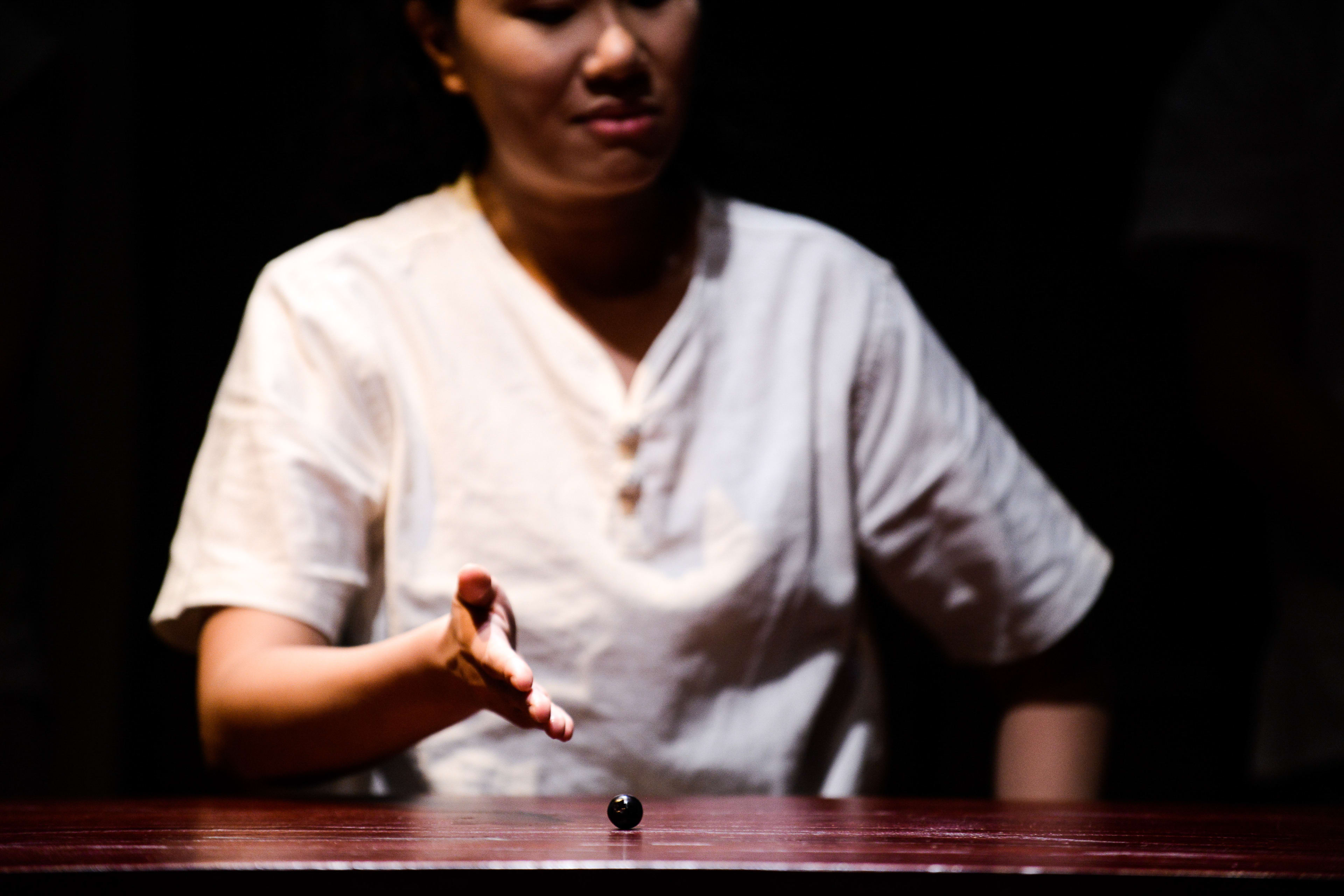 In a dramatic, dimly lit scene from Rooftop Productions’ contemporary theatre piece 'Milk and Honey', a performer in a simple white shirt reaches out with an open hand toward a single, solitary black marble centered on a wooden table. The stark lighting and minimalist staging emphasize a moment of quiet tension and introspection, characteristic of the company's experimental approach to storytelling in the Hong Kong stage scene. 'Milk and Honey'