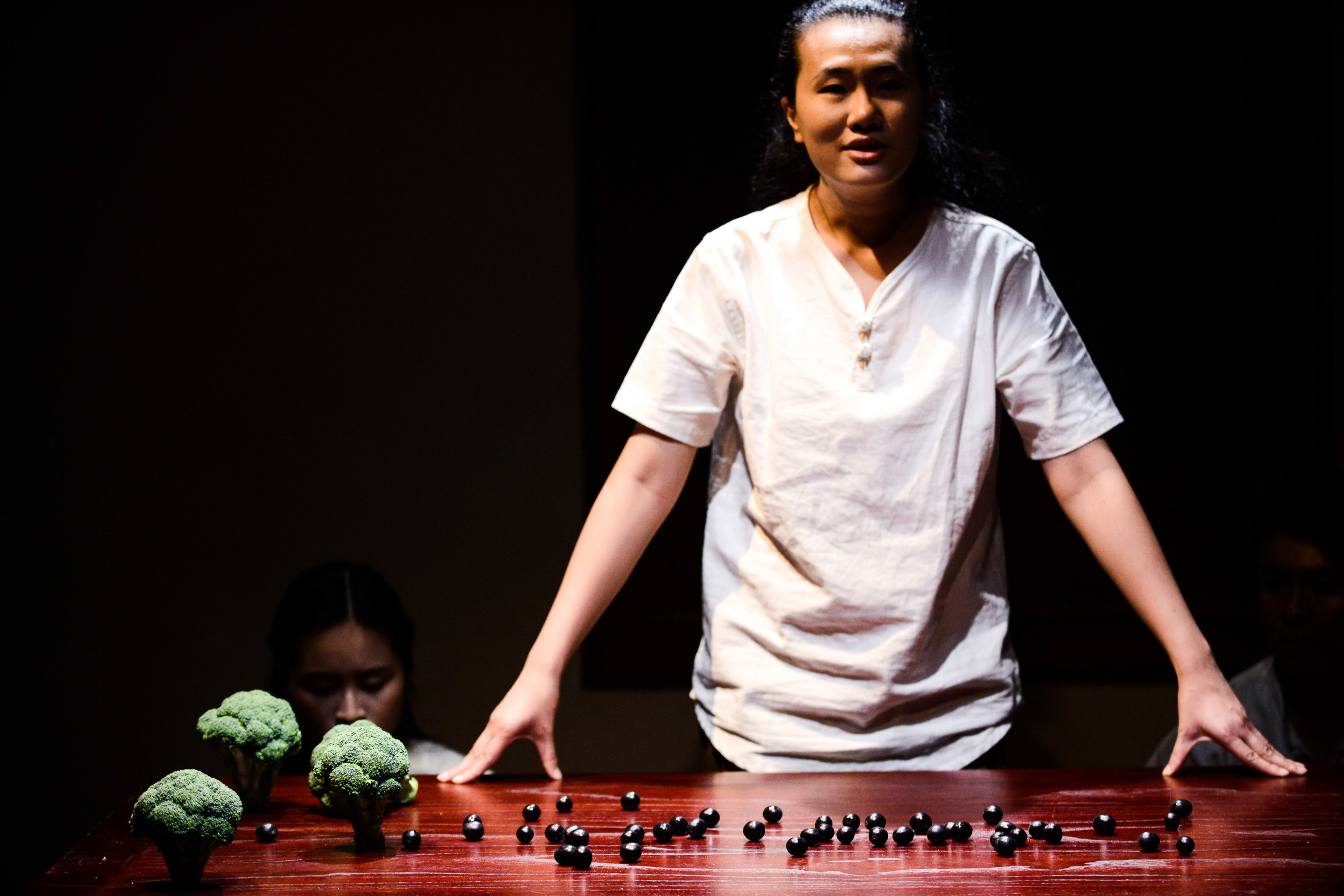 在天台製作 (Rooftop Productions) 的當代劇場作品《牛奶與蜜糖》中，演員李婉晶站在舞台中央，在黑暗的背景下被聚光燈照亮。她俯身在木桌前，桌上散落著深色的果實，兩株西蘭花像樹木一樣立在旁邊。演員隆藹宜在她的左側，身影隱約可見，靜靜觀察著台上的一切。這部作品透過極簡的舞台設計，探討親密關係與人際連結，正如劇名所寓意：『牛奶與蜜糖』。