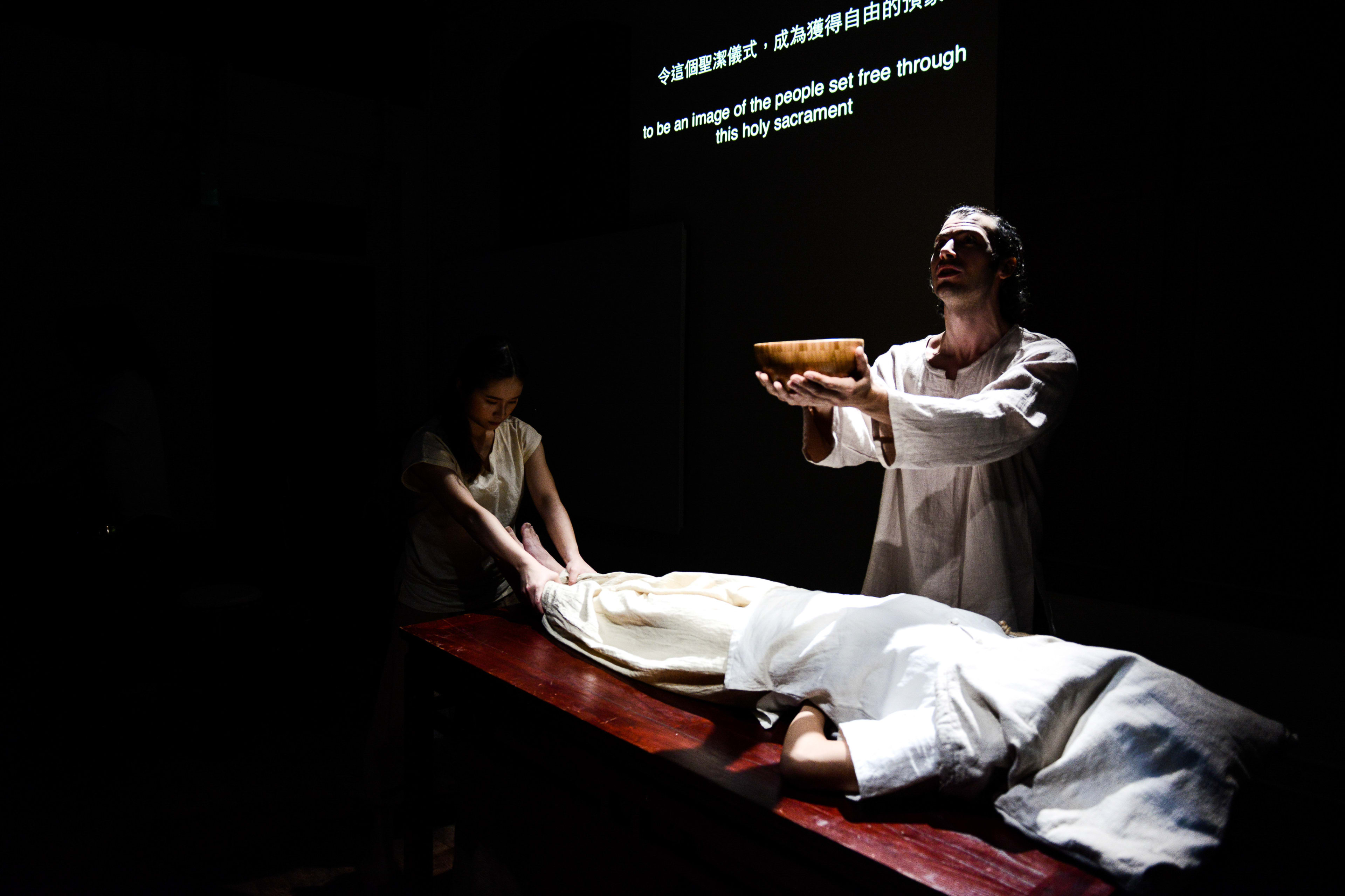在這張天台製作（Rooftop Productions）當代劇場作品《牛奶與蜜糖》的劇照中，演員艾浩家（Ivor Houlker）站在強烈的聚光燈下，神情莊嚴地雙手高舉一隻木碗。旁邊一位女性正照料著躺在木桌上、被白布覆蓋的角色。舞台四周漆黑，將觀眾的注意力聚焦於這場充滿儀式感的演出。背景投影著字幕：「令這個聖潔儀式，成為獲得自由的預像」。