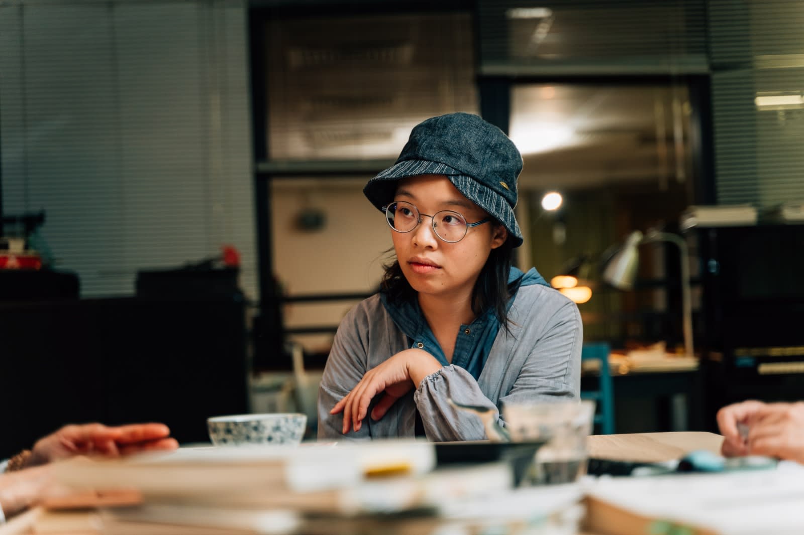 A close-up headshot of actor Miu Law captured during a theatre rehearsal for a Rooftop Productions (天台製作) contemporary stage performance. Miu is wearing a denim bucket hat and glasses, looking intently toward an off-screen collaborator with a thoughtful, focused expression, emphasizing the collaborative and intimate atmosphere of Hong Kong's independent theatre scene.