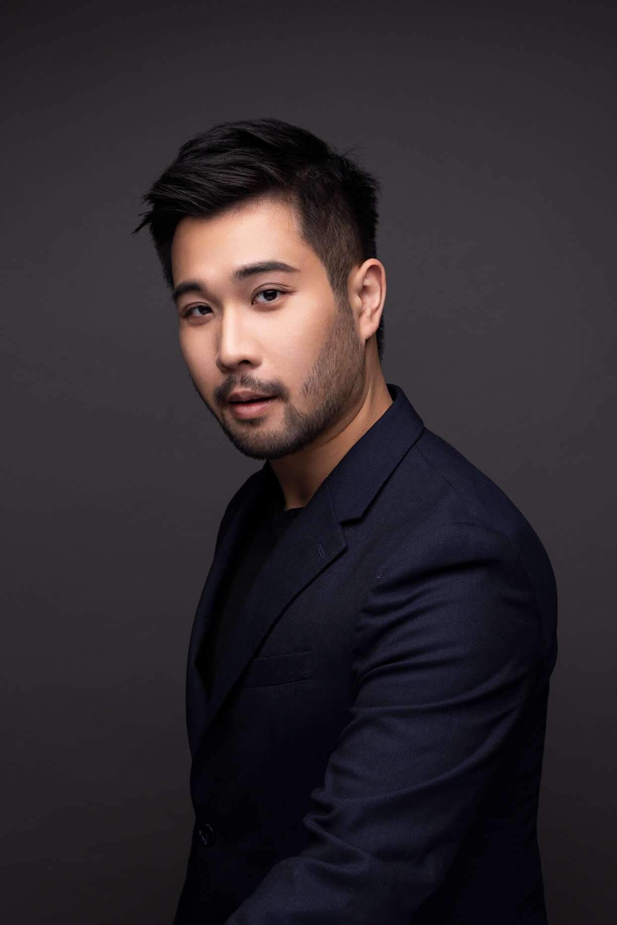 A professional headshot of actor Mok Kok-pong for Rooftop Productions (天台製作). He is captured in a medium close-up, turned slightly to the side against a dark, moody background, wearing a sharp navy blue blazer. His expression is focused and contemplative, reflecting the high standards of contemporary Hong Kong theatre performances.