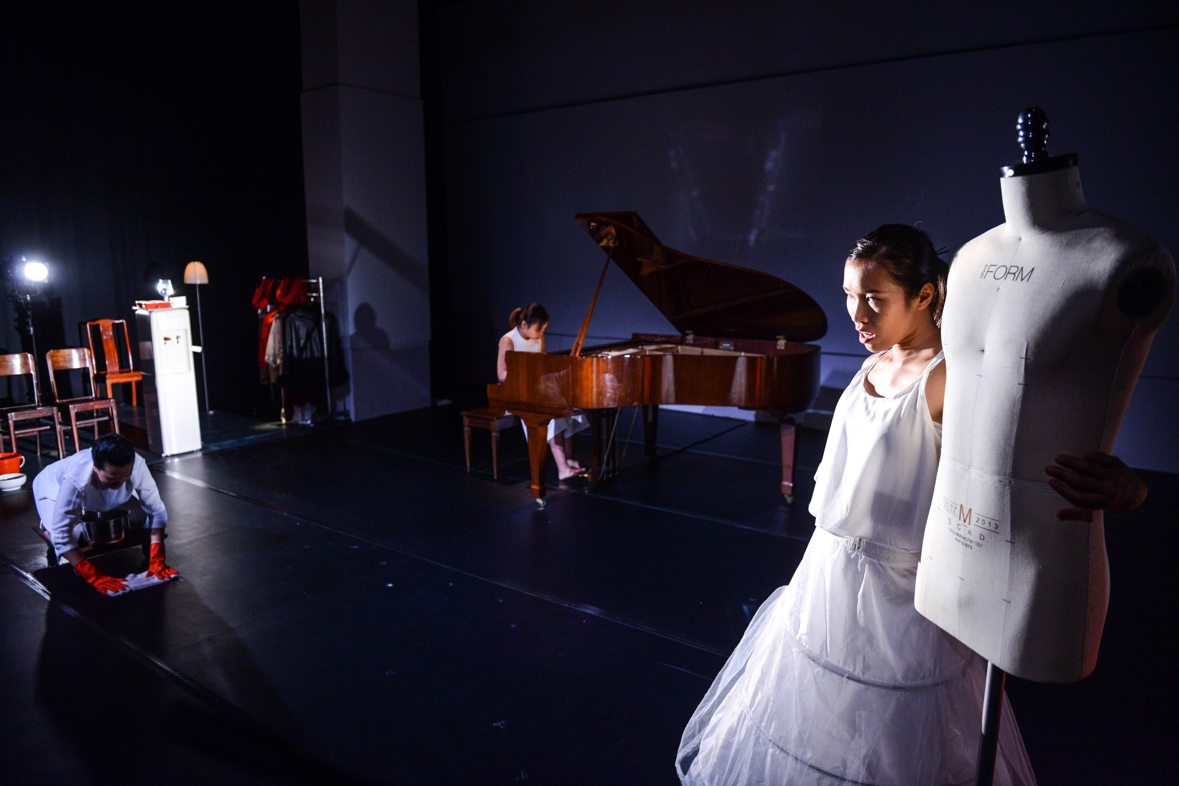 在這場由香港天台製作（Rooftop Productions）呈獻的當代劇場演出《不是女僕》（Not The Maids）中，三位表演者在昏暗簡約的舞台上呈現出關於勞動與身份的沉思。前景中，身穿白裙的表演者站在人台旁，神情若有所思；左側的表演者戴著紅手套正在擦拭地板，背景處則有一位表演者正坐於鋼琴前。透過這幅畫面，演員李婉晶低聲訴說著對塵埃的定義：「塵，有分乾淨嘅塵，同污糟嘅塵。」