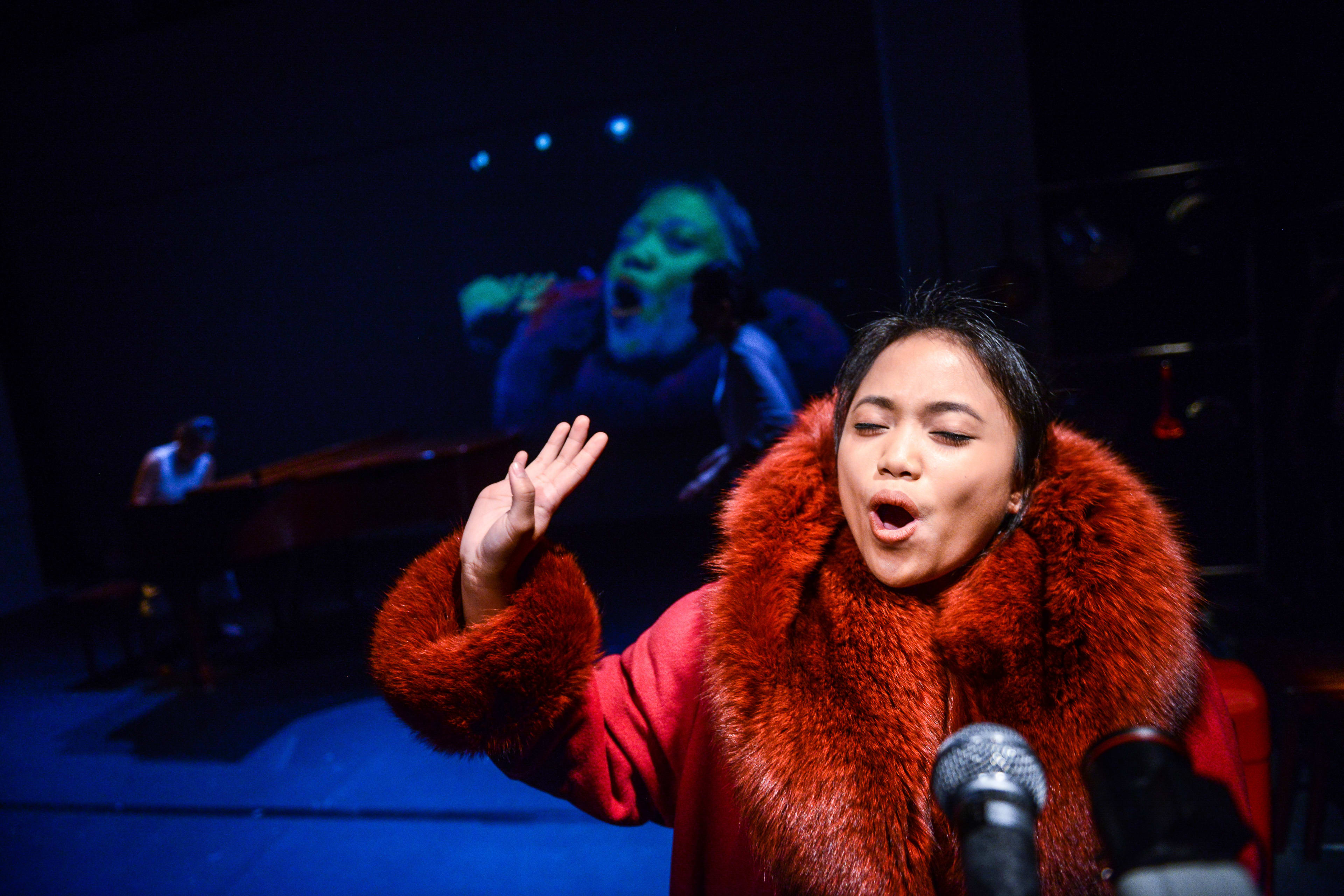 演員區洛思 (Russell Terre Aranza) 在天台製作 (Rooftop Productions) 的當代香港劇作《不是女僕》中登台演出。在冷調藍色燈光的映襯下，她身穿帶有誇張毛領的紅色大衣，神情專注且動作豐富，背景投射出一張巨大的變形人臉，營造出強烈的戲劇氛圍。區洛思在台上演繹道：「你們在做什麼？我看開會回來，而我期望所有東西都完美。」