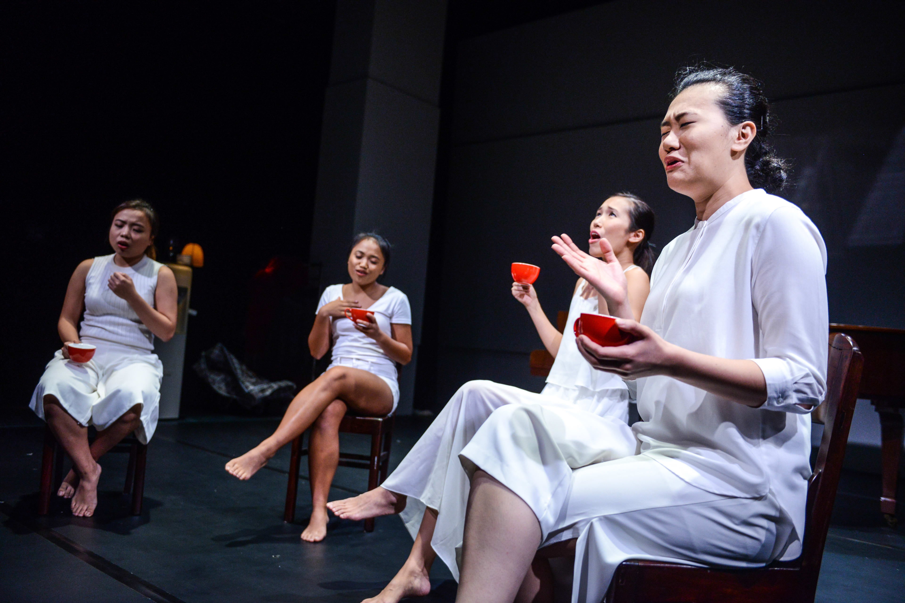 在天台製作 (Rooftop Productions) 當代香港劇場作品《不是女僕》(Not The Maids) 的排練場景中，四位演員身穿統一的白色服裝，坐在舞台簡約的木椅上。她們手中各持一個紅色小杯，表情交織著沉思與情感張力，深刻呈現出劇作對香港家庭傭工關係的探討。前景中一位演員正深情演繹台詞：「我屋企係我15歲左右開始請『工人姐姐』。」
